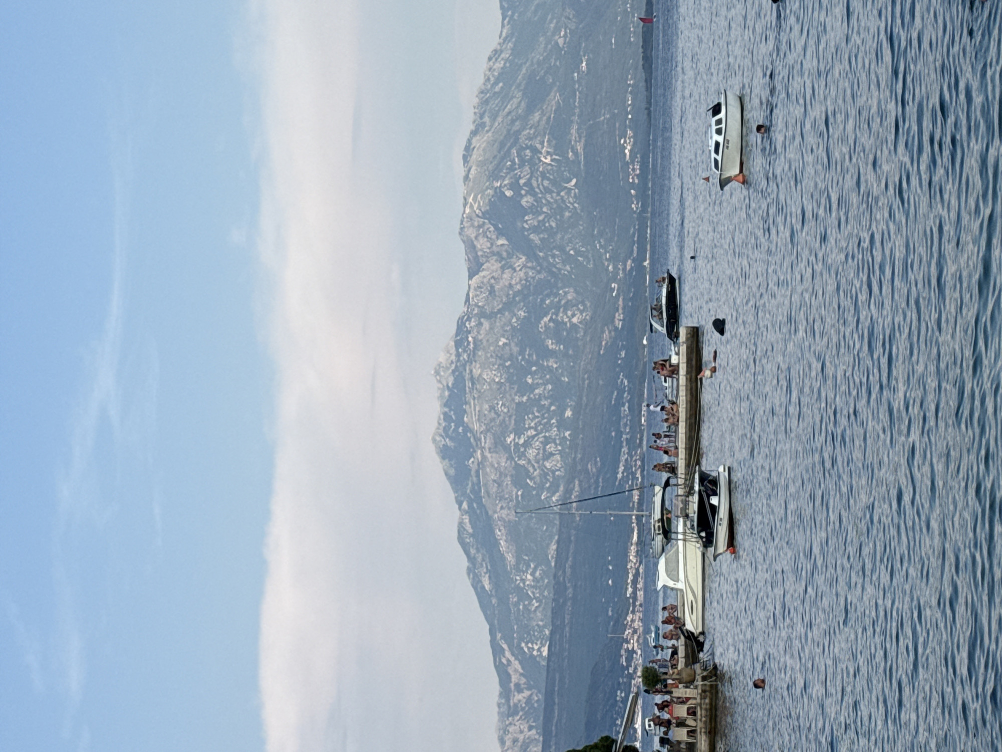 This media file shows a scenic view of a body of water with several boats docked and people enjoying leisure activities. In the foreground, a concrete pier lines the water's edge, with numerous individuals gathered, some seated and others standing. Various boats, including a sailboat and several motorboats, are moored to the pier. One motorboat is white with a dark hull, and another appears to be a smaller, open vessel. Further out in the water, a white boat with a dark roof is visible, with a person swimming nearby.
Dominating the background is a rugged, mountainous landscape. The slopes are covered in vegetation and show signs of rocky terrain. A town or village is nestled at the base of the mountains, with buildings clustered along the coastline.
The time of day appears to be daytime, possibly late afternoon, given the soft lighting and the angle of the sun. The weather seems pleasant, with a clear blue sky and some wispy white clouds. The water is calm with gentle ripples, reflecting the light. The overall atmosphere suggests a relaxed holiday or vacation setting. There is no visible text in the media file. The location context provided is Đenovići, Montenegro.