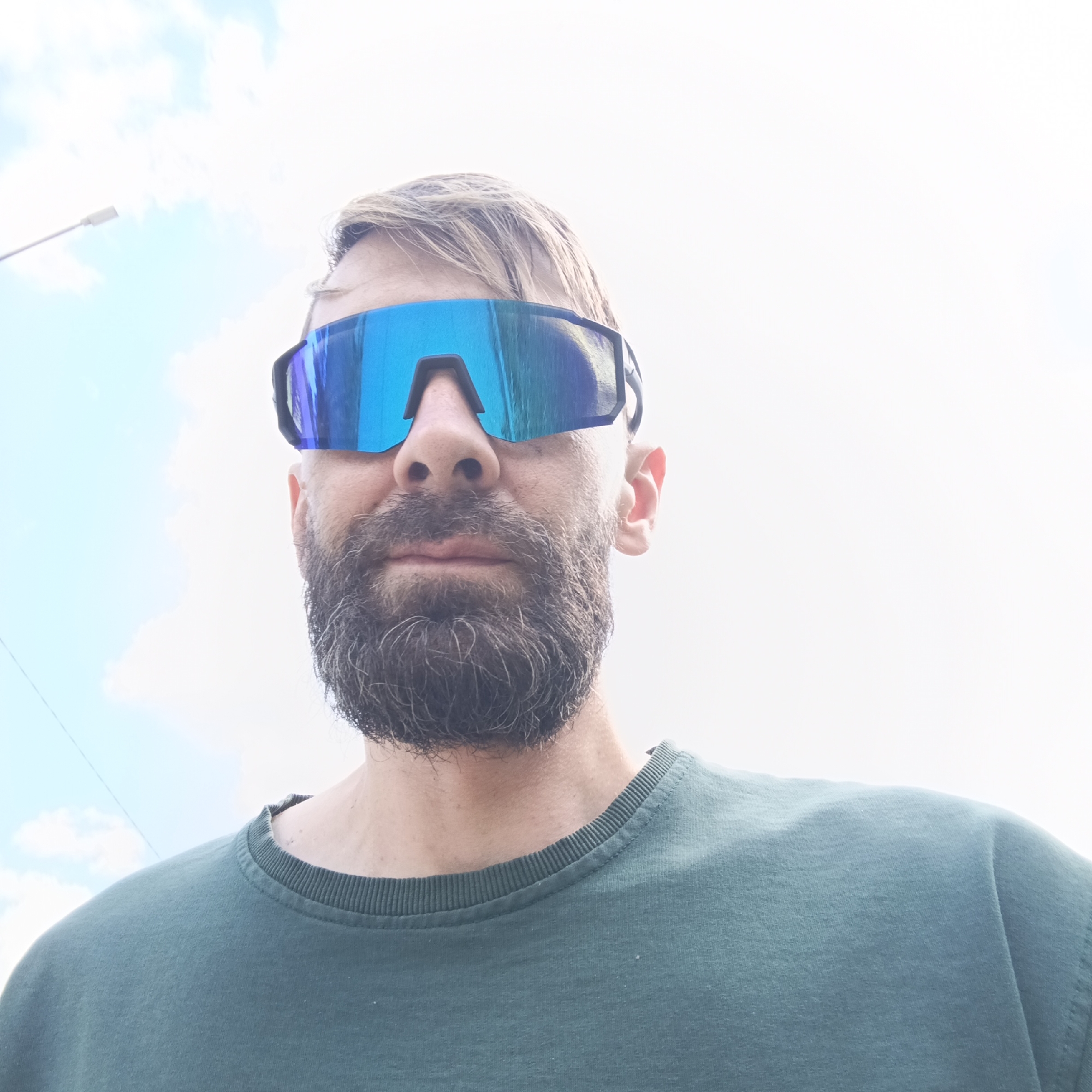 The image shows a close-up, low-angle, outdoor, waist-up shot of a Caucasian man wearing large, blue-tinted, wraparound sunglasses. He has a beard and short, light brown hair. He is wearing a dark green crew-neck sweatshirt. The background is a bright, overcast sky with some faint blue hues and wisps of clouds. A thin wire or cable runs diagonally across the upper left portion of the frame. The location is identified as Марковский, Russia.