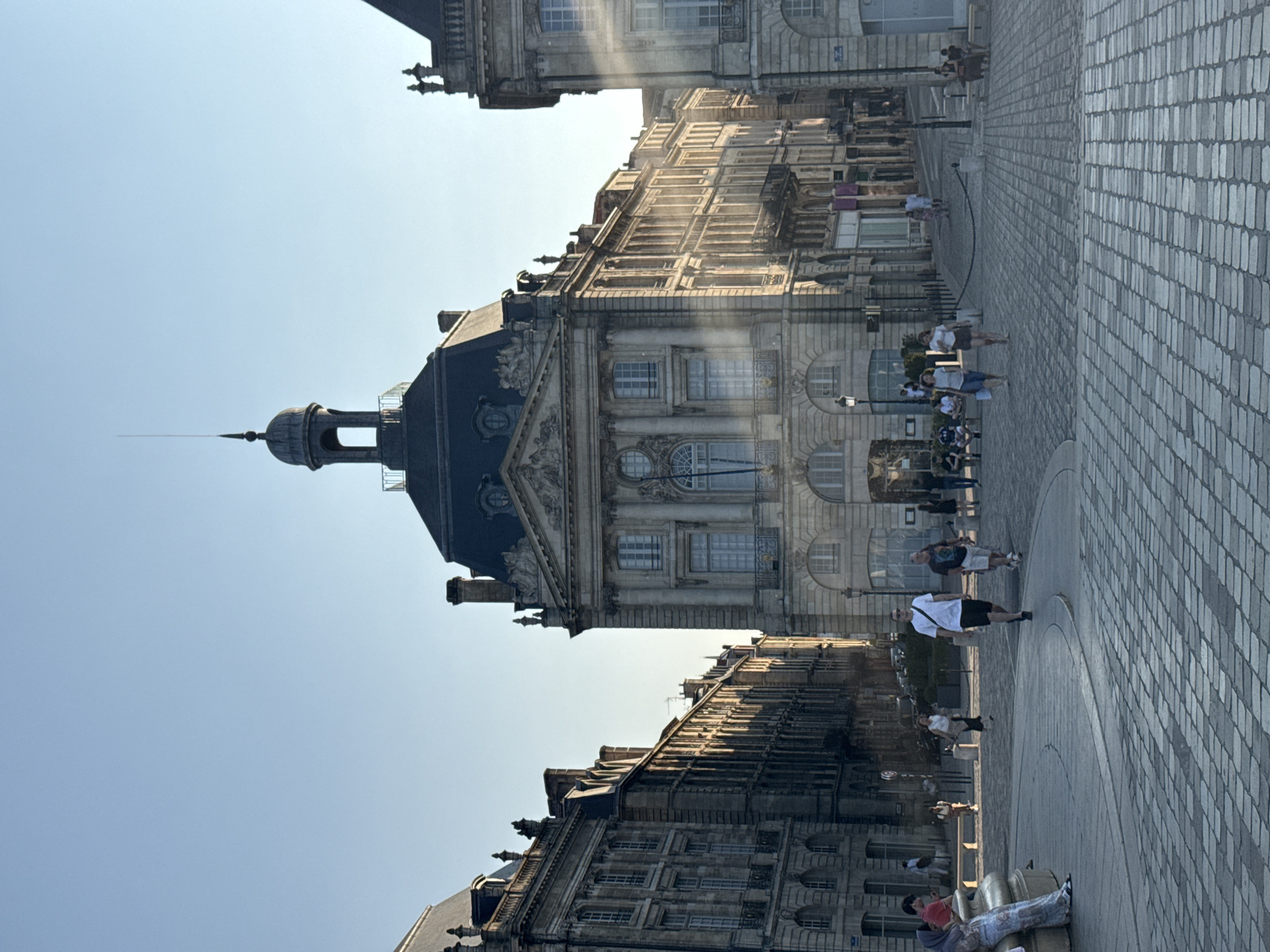 The image depicts a sunlit public square in Bordeaux, France, likely Place de la Bourse, given the architectural style of the surrounding buildings. The foreground is paved with cobblestones, curving slightly. In the midground, several people are walking and standing. A man in a white t-shirt and black shorts is walking towards the viewer, while other individuals are seated on benches or strolling. The background features grand stone buildings with classical architectural elements, including arched windows and ornate facades. A prominent building in the center has a domed cupola topped with a spire. Sunlight casts long shadows, indicating it is either early morning or late afternoon. The sky is clear and blue.