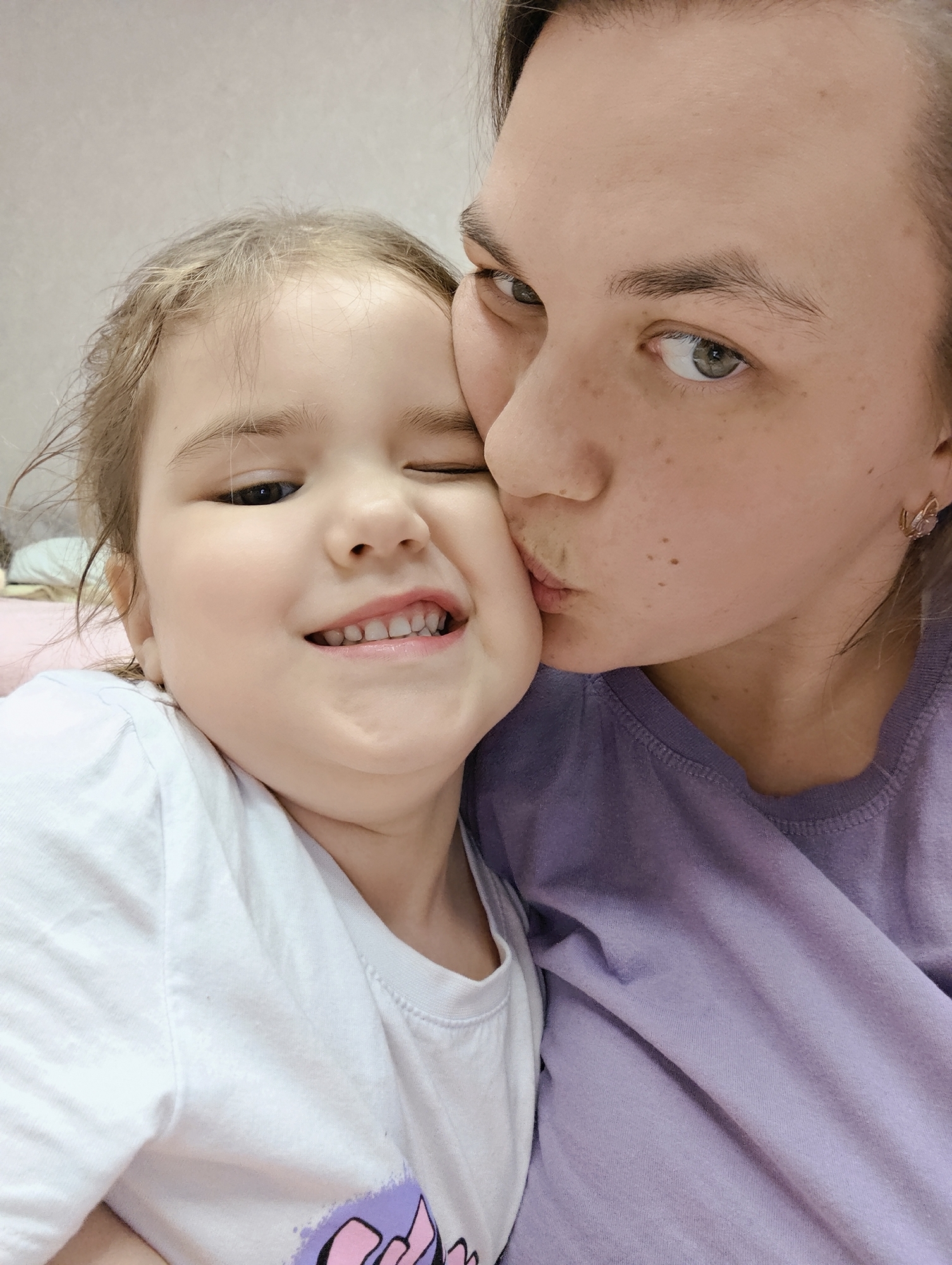 A close-up shot shows a young girl with light brown hair, wearing a white t-shirt with a purple and pink design, smiling and winking. A woman with light skin and short brown hair is kissing the girl's cheek. She wears a light purple shirt and has butterfly earrings. The background is a light beige wall. The location is Chaikovsky, Russia.