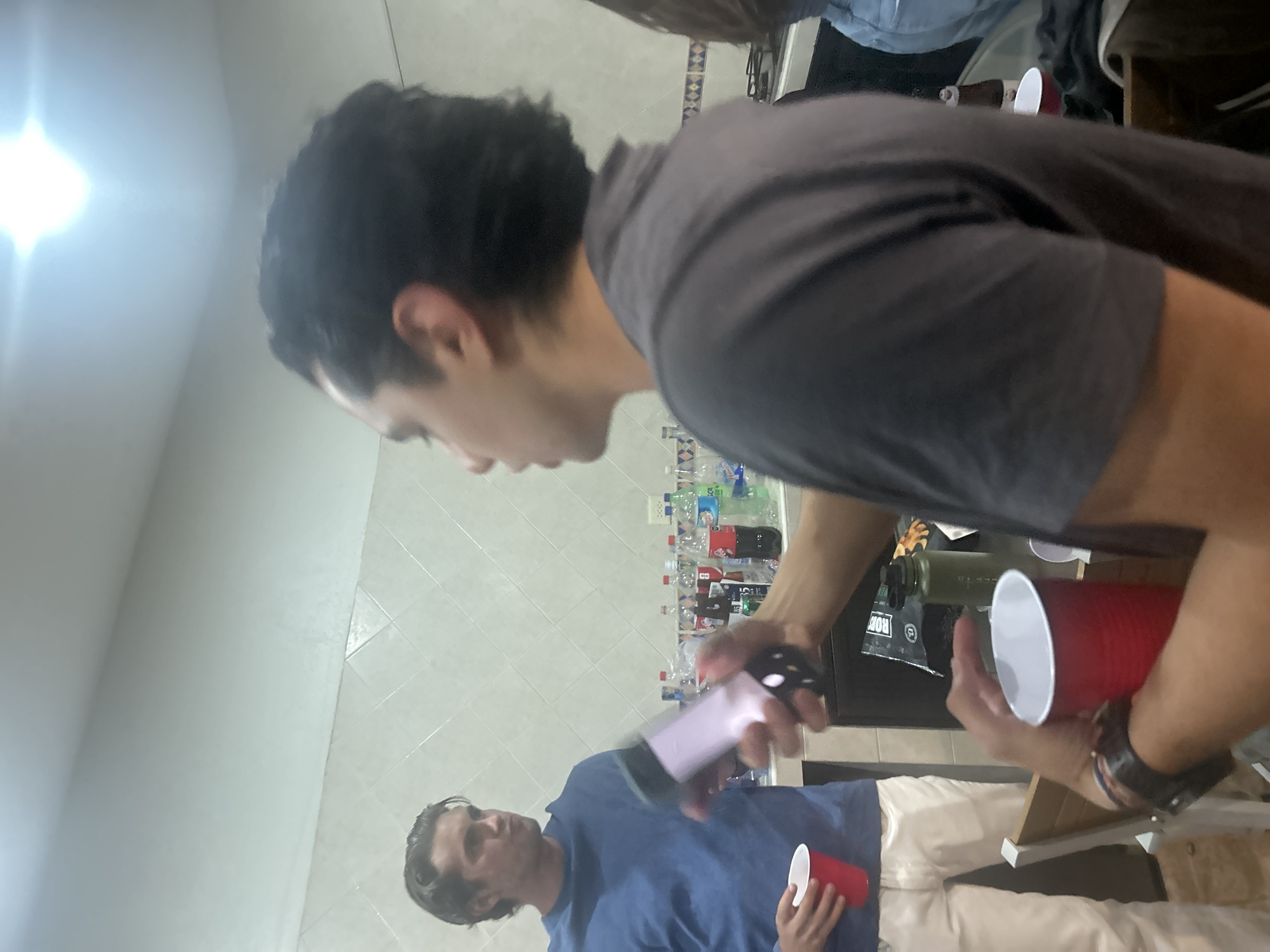 A group of people are indoors, likely at a social gathering. One man is in the foreground, wearing a grey shirt and holding a red plastic cup. Another man is holding a phone with its camera activated in the direction of the man with the cup. He is wearing a blue shirt and white pants, and also holding a red cup. The background has a tiled wall with a countertop covered with water bottles and other objects. It is unclear where in San Juan Cuautlancingo, Mexico this image was taken.