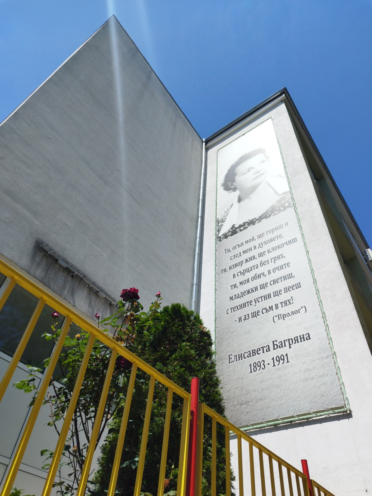 The image shows a building exterior in Sofia, Bulgaria, featuring a large banner of Elisaveta Bagriana, a Bulgarian poet. The banner includes a black and white portrait of Bagriana, along with some of her poetry and her birth and death years (1893-1991). In the foreground, there is a yellow metal fence and some greenery, including a rose bush with dark red roses. The building is light grey and appears to be made of concrete. The sky is blue and mostly clear. The shot is taken from a low angle, looking upwards towards the building and the banner.