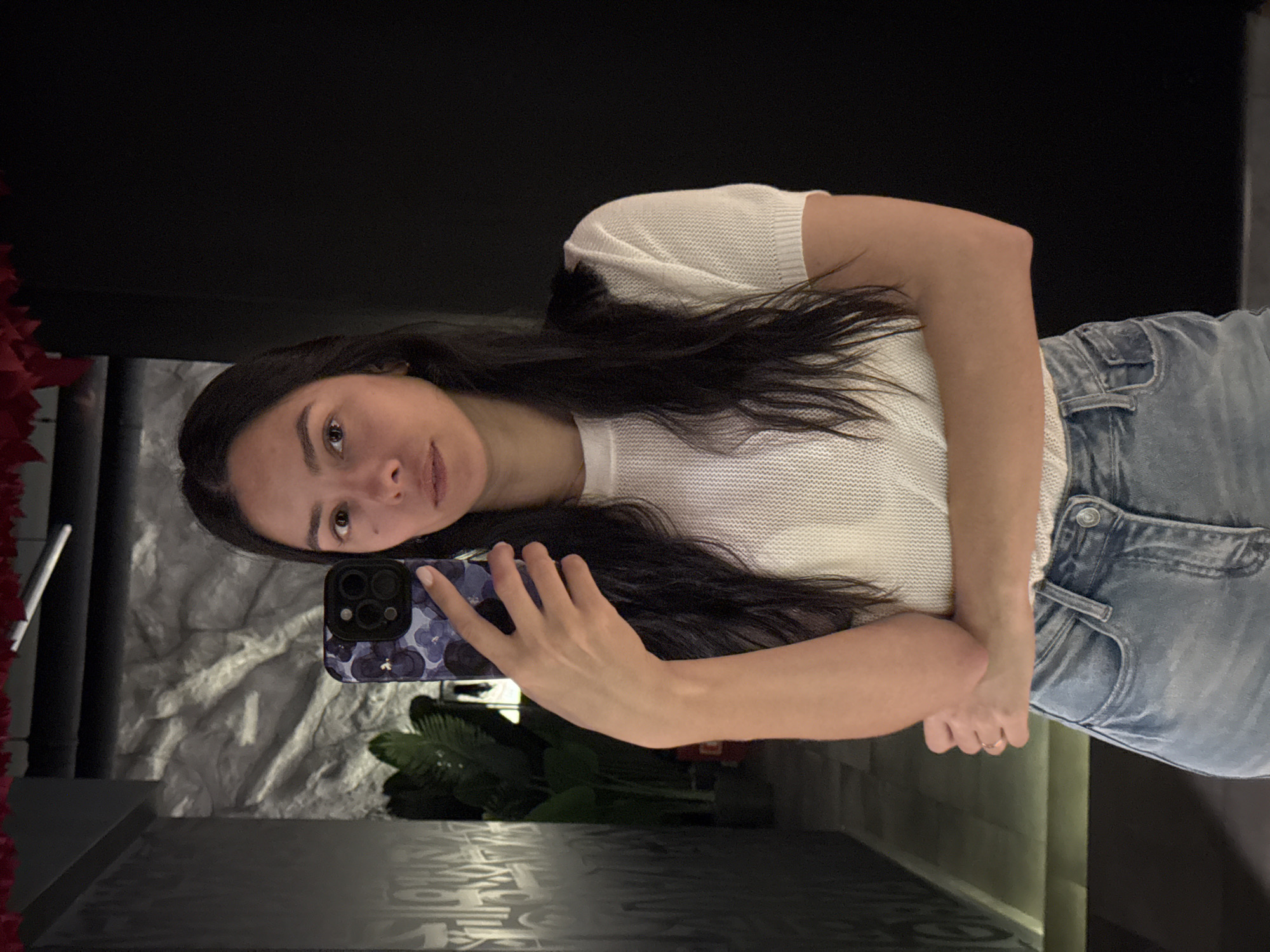 A woman is taking a selfie in a room that appears to be a bathroom, as evidenced by the mirror and the surrounding environment. She is holding a mobile phone with a patterned case. She is wearing a white short-sleeved top and light blue jeans. The background includes textured walls, a plant, and a dark surface below the mirror. The scene appears to be indoors, possibly in a private residence. There is no information in the image to indicate this location is in Chaikovsky, Russia.