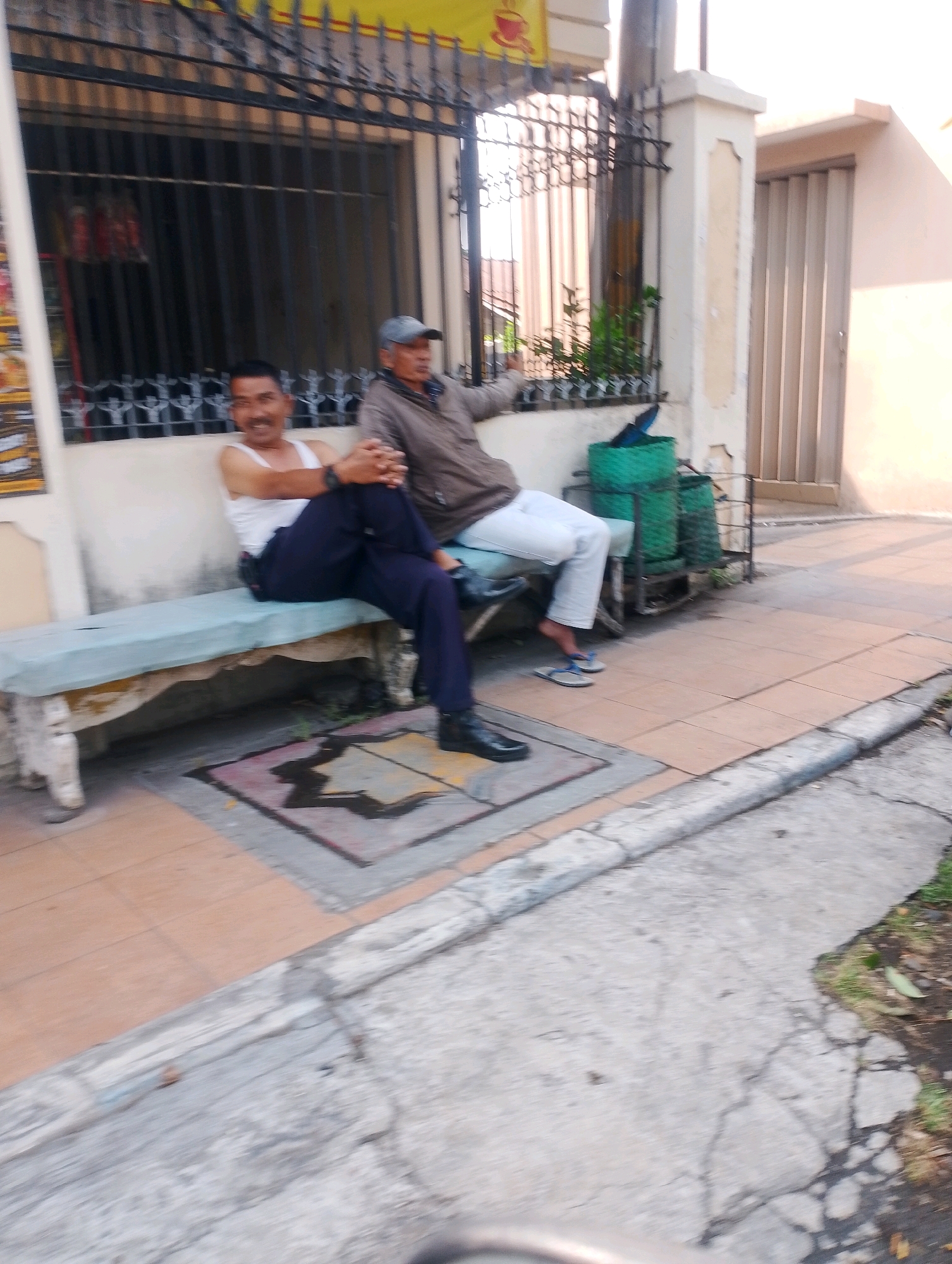 Two men are sitting on a bench in front of a building with barred windows. The man on the left is wearing a white tank top and dark pants, and is smiling at the camera. The man on the right is wearing a brown jacket and a blue cap, and is looking away from the camera. There are two green trash cans behind the bench. The sidewalk in front of the bench is tiled. The scene appears to be in an urban area, possibly in Probolinggo, Indonesia, given the architectural style of the building and the presence of outdoor seating.