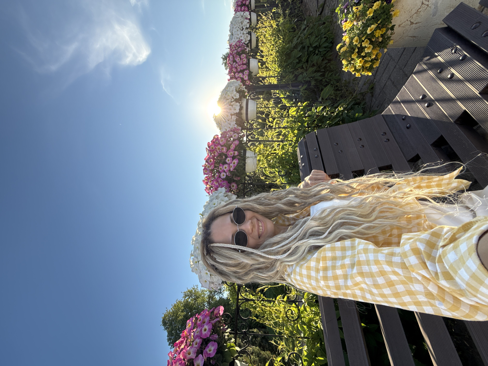The image captures a smiling young woman seated on a dark grey slatted bench in an outdoor setting during golden hour. She has long, wavy blonde hair with visible braids and wears dark oval sunglasses, a yellow and white gingham check shirt, and a white top. Behind her, an ornate black metal fence supports a linear arrangement of white hanging planters filled with pink/purple and white petunias. Lush green foliage is visible beneath and behind the fence. The sun, emanating from behind one of the central white flower baskets, creates a prominent starburst effect against the clear blue sky, which features some wispy clouds. To the right, a stone pot containing yellow flowers is situated on a paved surface. The lighting indicates strong, low-angle sunlight, casting long shadows. This scene is located at the Volga Embankment in Yaroslavl, Russia, a public area known for its decorative floral displays.