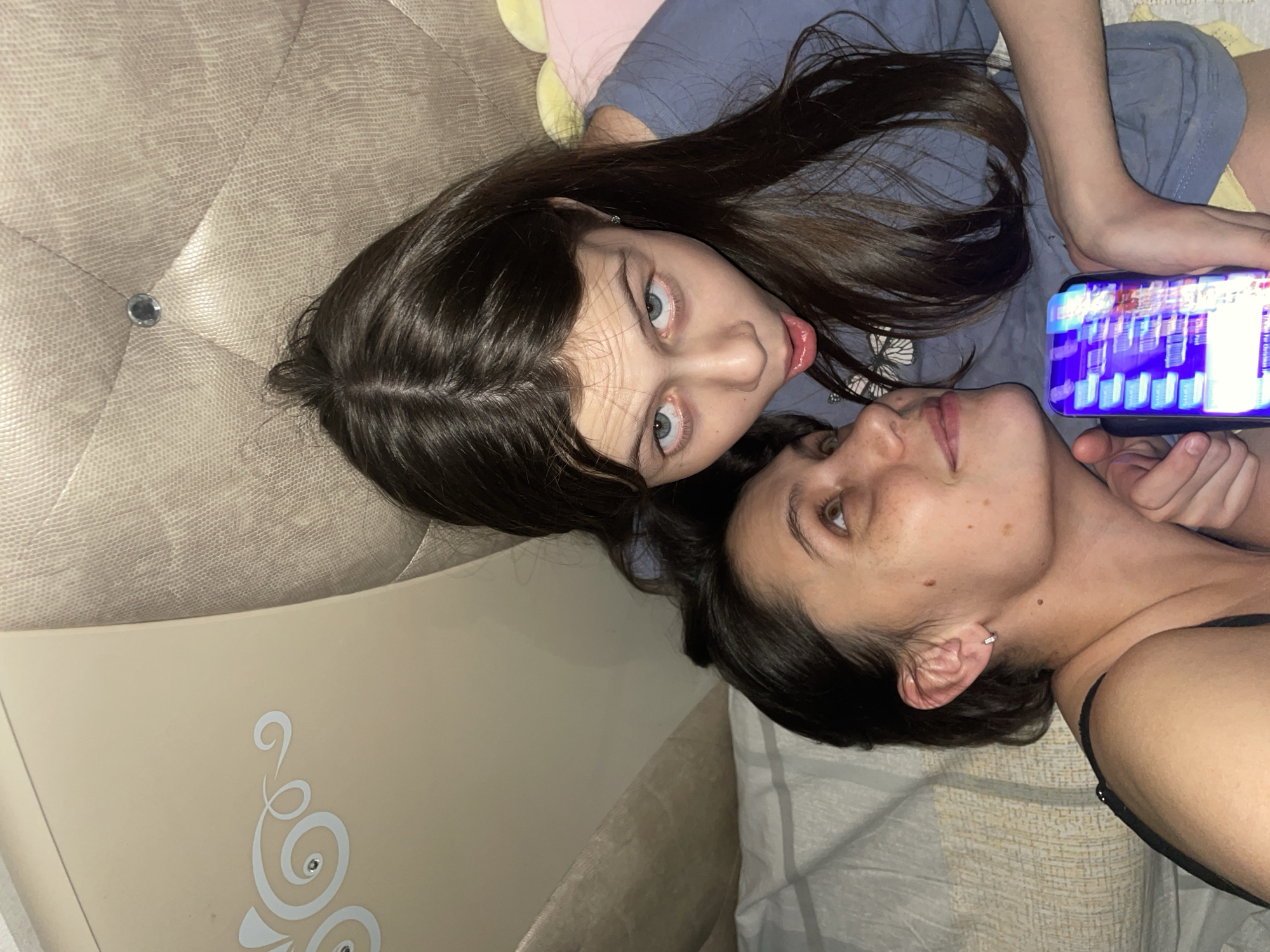 Two individuals, likely female, are captured in a close-up, angled view. The younger of the two has long, dark brown hair, fair skin, and prominent, light-colored eyes. She is wearing a bluish-gray shirt and looking directly at the camera. Her mouth is slightly open, revealing a portion of her tongue. The other person, presumably an adult female, has darker features, shorter dark hair, and appears to be reclining with her head near the younger person's. She is holding a brightly lit smartphone, potentially displaying an image or video. The background consists of beige-colored bedding, possibly a bed or couch. The location cannot be determined from the image. There is nothing in the image to indicate that it was taken in the city of Izhevsk, Russia.
