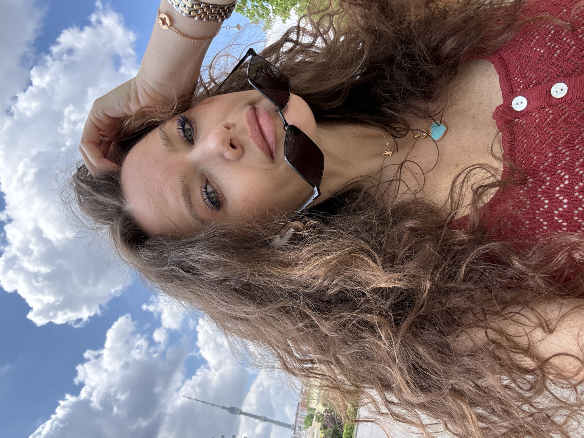 A woman with long, curly brown hair is the central subject of the image. She is wearing sunglasses perched on her nose and a maroon top with white buttons. A gold necklace with a light blue pendant adorns her neck. Her right hand, adorned with bracelets, rests on her head.
The background reveals a cityscape under a partly cloudy blue sky. Distinctly visible is the Ostankino Tower, a prominent television and radio tower in Moscow, Russia. The environment suggests the woman is outdoors in a location with a clear view of the Moscow skyline.