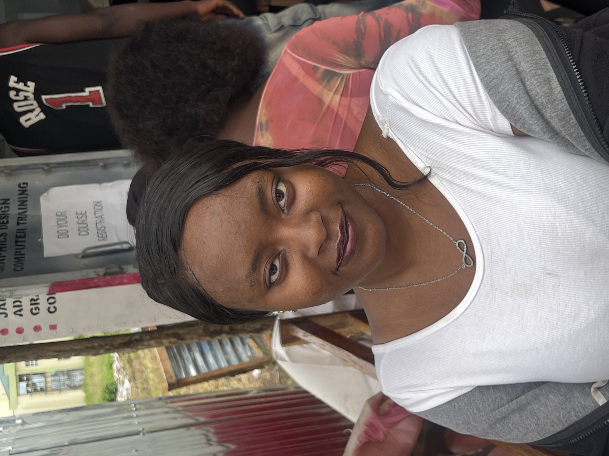 This is a medium-close-up, eye-level portrait of a young Black woman. She is facing the camera, with her head slightly tilted to her right. Her skin is dark, and she has dark hair that is pulled back from her face, with some strands falling to her left side. She is wearing a white V-neck shirt with grey sleeves and a black zipper jacket. Around her neck, she wears a silver necklace with an infinity symbol pendant. Her makeup appears to include dark lipstick.
Behind her, slightly out of focus, are other individuals and elements of an outdoor environment. To her left, there is a partially visible structure made of corrugated metal. To her right, a person in a black basketball jersey with the name "ROSE" and the number "1" is visible. Further back, another person is wearing a pink tie-dye garment. In the background, a building with light-colored walls and windows can be seen, surrounded by green foliage. A sign on a vertical surface behind the woman lists courses like "GRAPHICS DESIGN," "COMPUTER TRAINING," "AD," "GR," and "CO." The scene suggests an outdoor setting in a semi-urban or commercial area, possibly a training center or market stall. The location is identified as Wumat, Nigeria.