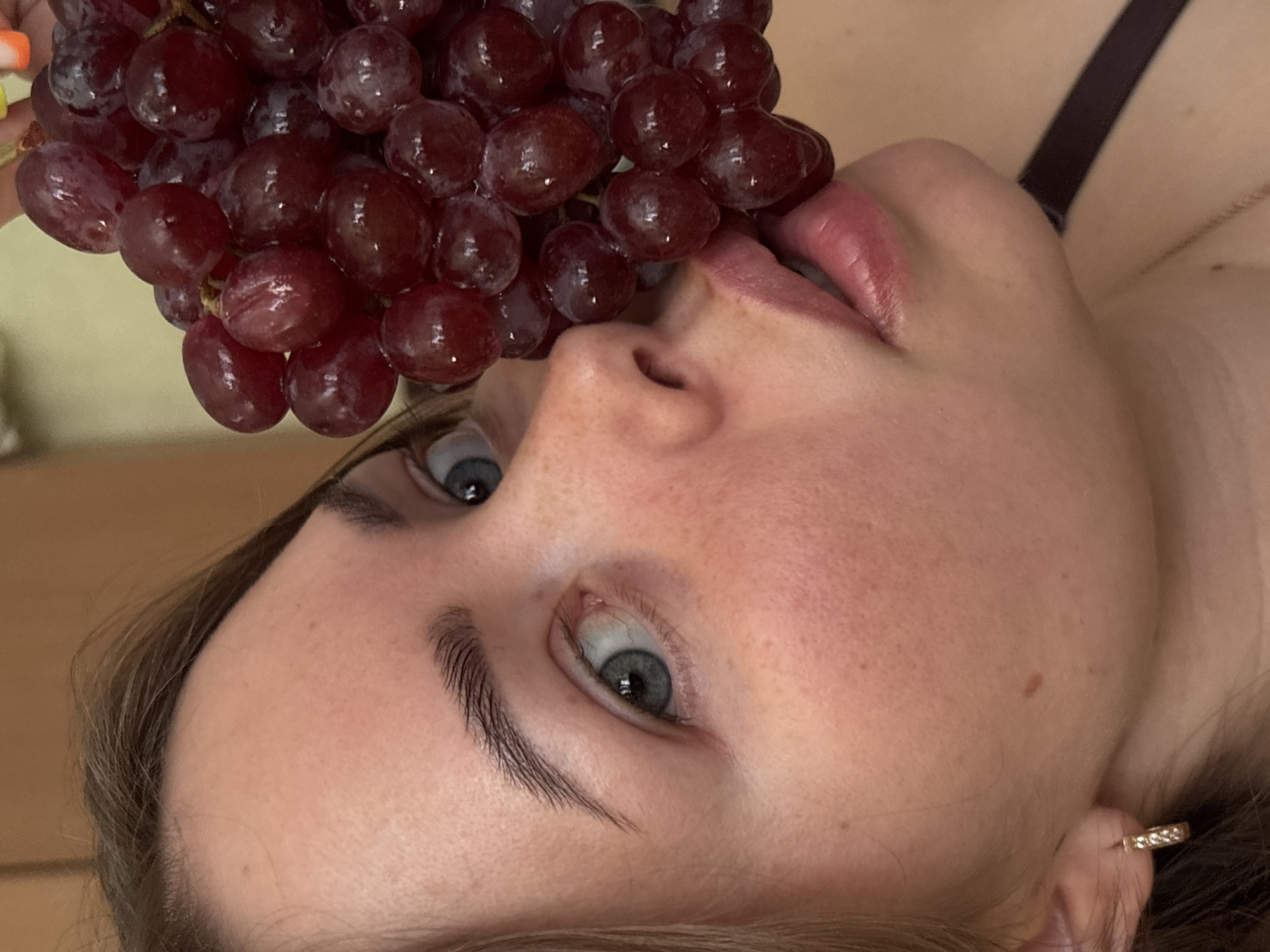 The close-up, top-down photograph, taken in Izhevsk, Russia, features a person holding a bunch of red grapes near their mouth. The person's face is partially visible, angled and tilted, with their blue-grey eyes looking upwards and slightly to the left. Their lips are slightly parted, touching one of the grapes, suggesting they are about to eat or are in the process of eating the fruit.
The subject has visible eyelashes and well-defined eyebrows. Their skin appears natural, showing some freckles. Light brown hair frames the face, and a small golden hoop earring, adorned with tiny stones, is visible in their right ear. A dark strap, possibly from clothing, is visible in the upper right background.
The bunch of grapes is prominent in the foreground, filling a significant portion of the image. The grapes are plump, firm, and deep red or purplish in color, with some appearing to have a natural bloom on their skin. The background is softly blurred, showing indistinct light yellowish-beige tones, indicating an indoor setting. The overall impression is one of natural light, capturing a moment of relaxed indulgence or enjoyment. There is no visible text.