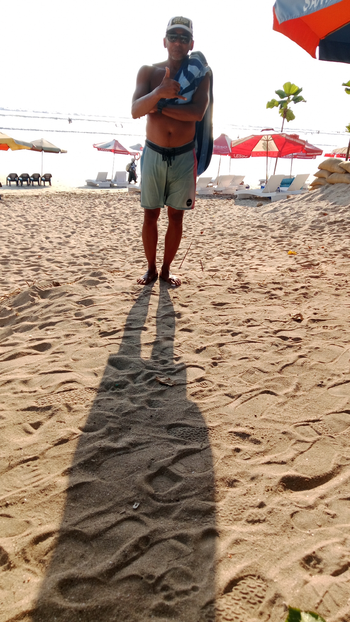A shirtless man wearing a baseball cap, sunglasses, and swim trunks stands on a sandy beach, holding a towel over his shoulder and giving a thumbs-up gesture. The sun is bright, casting a long shadow of the man onto the sand. In the background, there are beach umbrellas, lounge chairs, and sandbags. The ocean is visible in the distance.