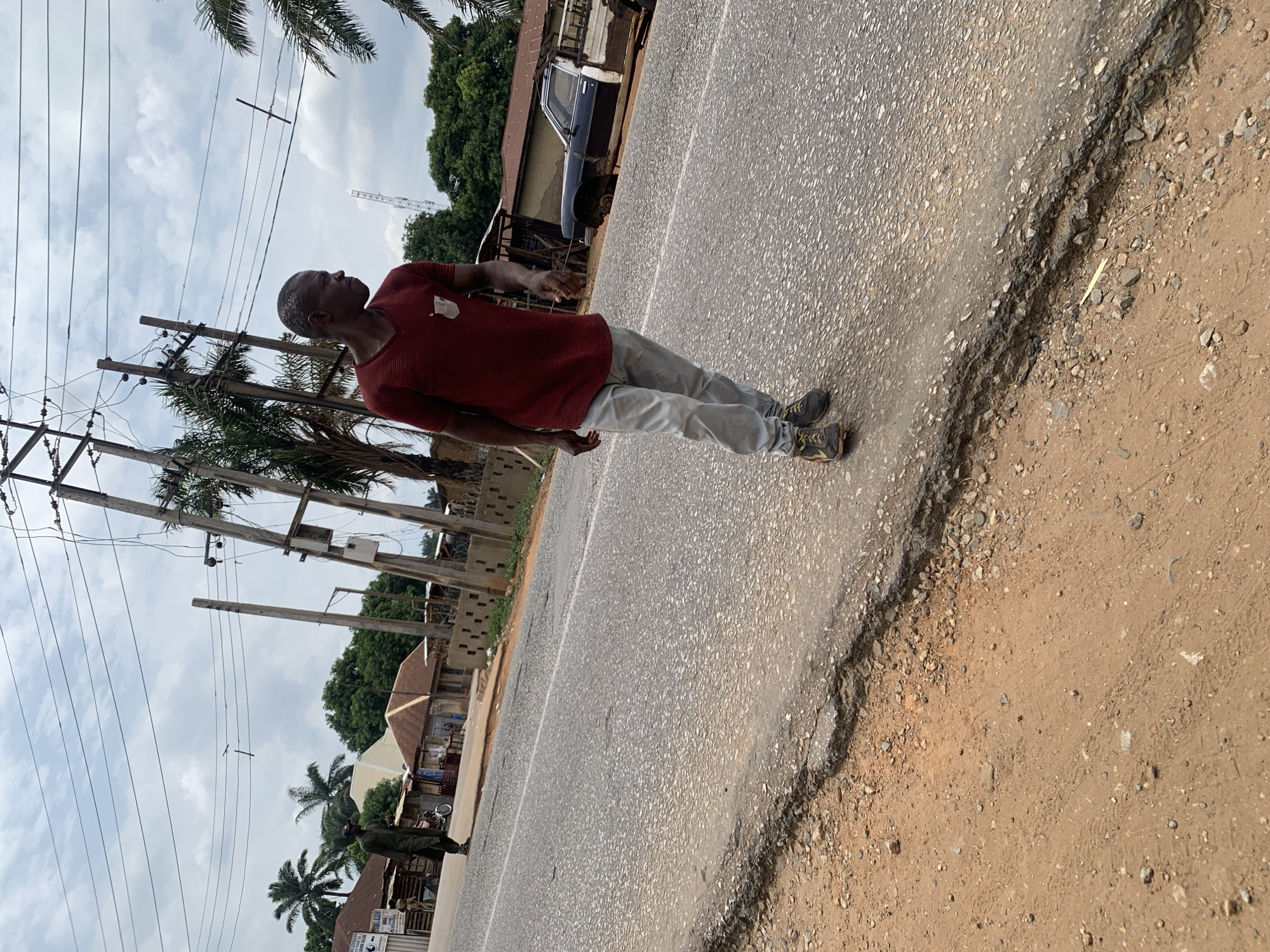 The image depicts an older man in a red shirt and light-colored pants standing on a cracked asphalt road. He is looking upwards and gesturing with his right hand. In the background, there are utility poles, palm trees, and buildings, suggesting an urban environment. A person in what appears to be a military uniform stands further down the road. Based on the prompt, the location is Akwanga, Nigeria. A derelict vehicle can be seen by one of the buildings.