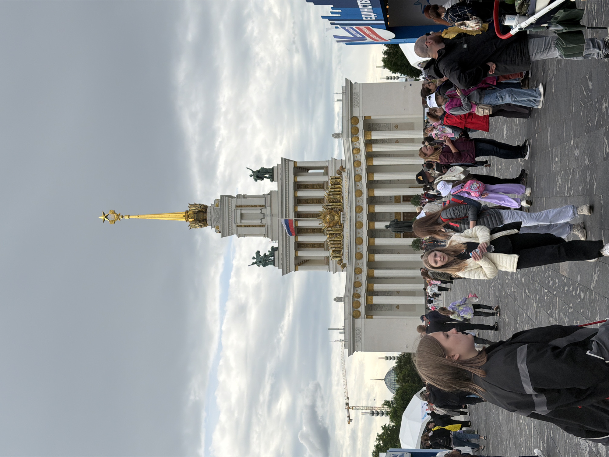 The image depicts the main pavilion of the All-Russia Exhibition Center (VDNKh) in Moscow, Russia. The pavilion, with its prominent golden spire topped with a star and its white columns, is the focal point of the image. A group of people is gathered in the foreground, likely tourists or visitors to the exhibition center. They are standing on a paved area, with some looking towards the pavilion. The sky is overcast, suggesting cloudy weather. In the background, a construction crane is visible, indicating ongoing work in the area. The image captures a typical scene at a popular tourist destination in Moscow.