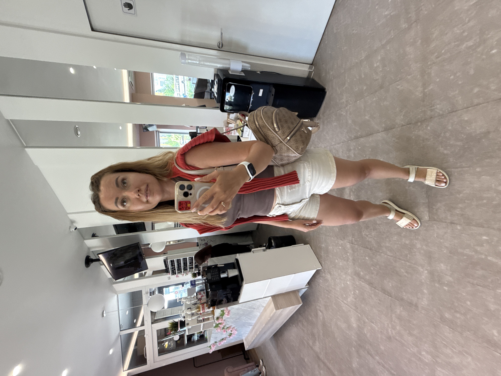 A blonde woman is standing in what appears to be a salon or waiting area. She is wearing a brown tank top, white denim shorts, a red sweater draped over her shoulders, and white sandals. She is holding a smartphone in her right hand, taking a selfie in a mirror. She has a small beige backpack with a logo on her left shoulder. The background shows a reception desk with a coffee machine, some plants, and shelves with bottles. There are mirrors on the wall and a water cooler. The floor is tiled.