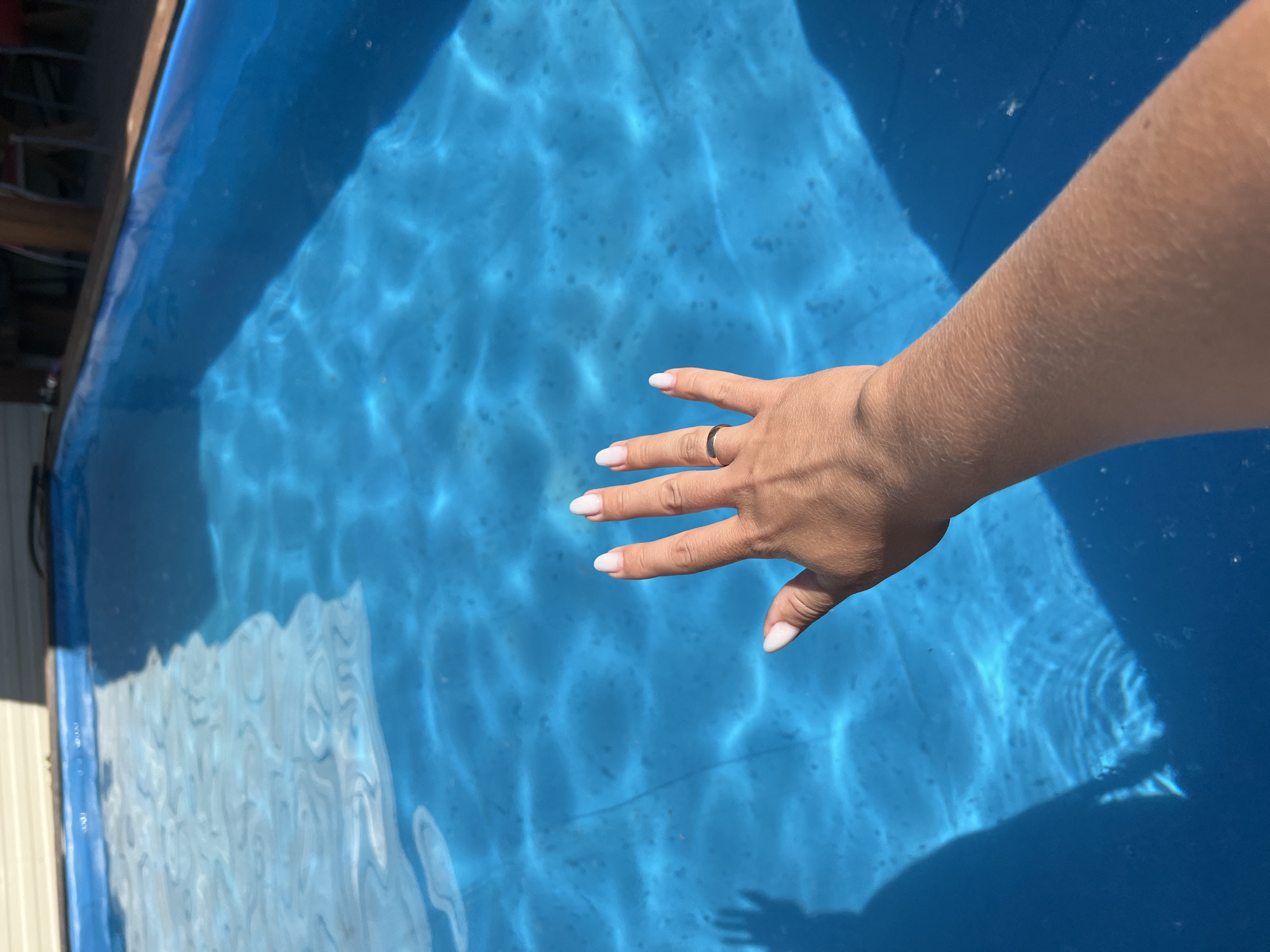 A first-person perspective shot shows a woman's left hand extended over a bright blue swimming pool. The hand has light-colored nail polish and a thin ring on the ring finger. Sunlight casts a shadow of the hand and arm on the water. The pool has visible ripples and is surrounded by a light-colored deck and some blurry background elements, including what appear to be chairs and a table. The image was taken outdoors on a sunny day. There is no information to suggest this location is in Novosibirsk, Russia.