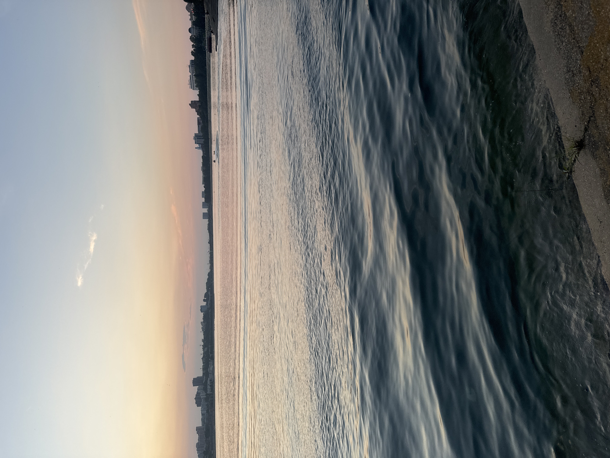 The image depicts a tranquil waterfront scene at dusk, likely along the Angara River in Irkutsk, Russia. The sky transitions from a pale blue at the top to soft hues of pink and orange near the horizon, indicating the sunset. A faint cloud is visible in the upper left portion of the sky.
A city skyline is silhouetted against the horizon, characterized by buildings of varying heights. On the water, there are a few small figures, possibly people on a jet ski, visible in the distance. The water itself is calm, with gentle ripples reflecting the colors of the sky. The foreground shows a concrete embankment or shoreline, with some sparse vegetation growing near the edge. The overall impression is one of peacefulness and the end of a day in a city situated by a large body of water.