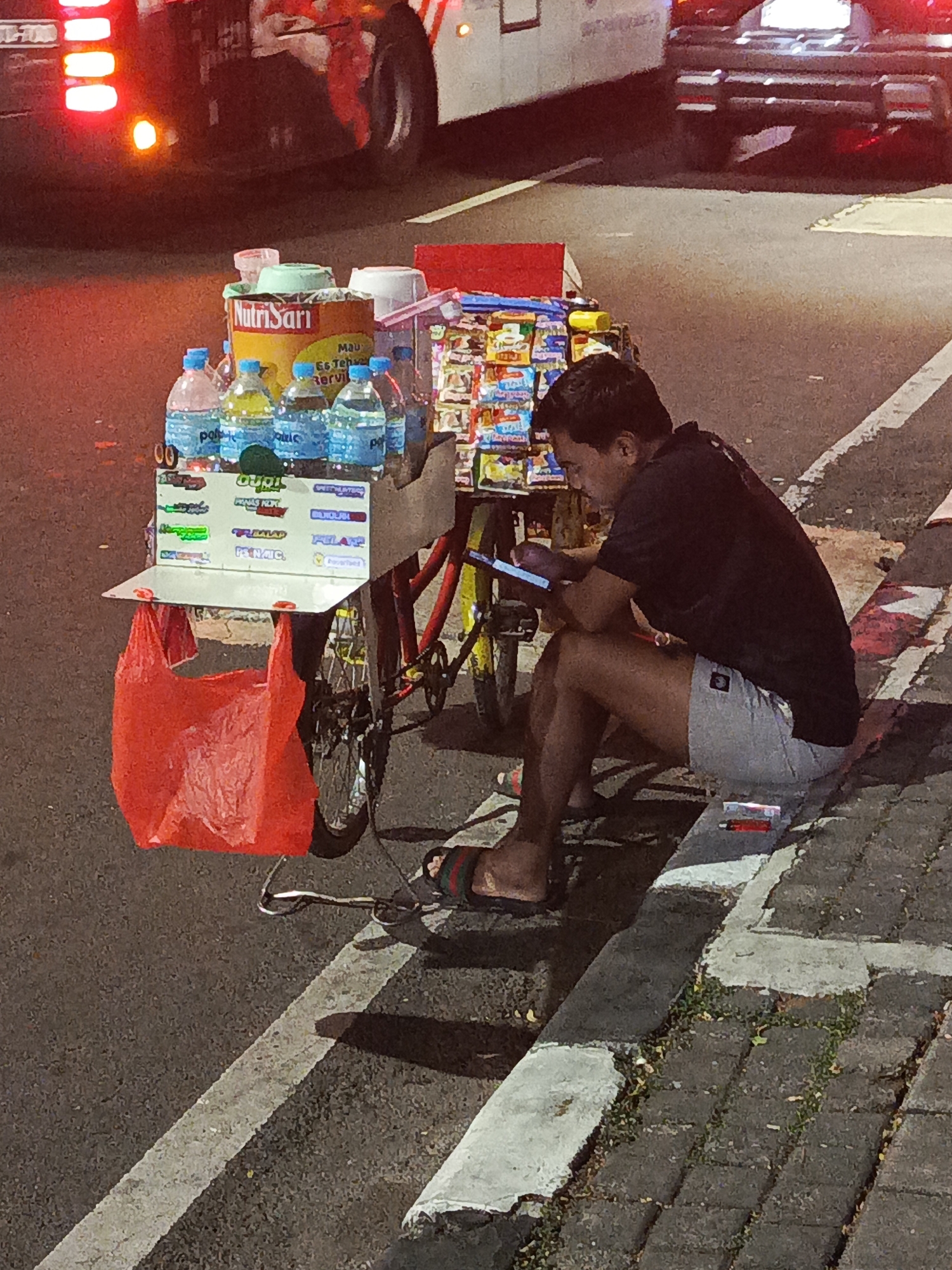 A man is sitting on the curb with his back to a street cart filled with drinks and snacks. He is looking at his phone. A large red plastic bag is hanging from the cart. In the background, a bus is visible. The scene appears to be in Central Jakarta, Indonesia, based on the context provided.