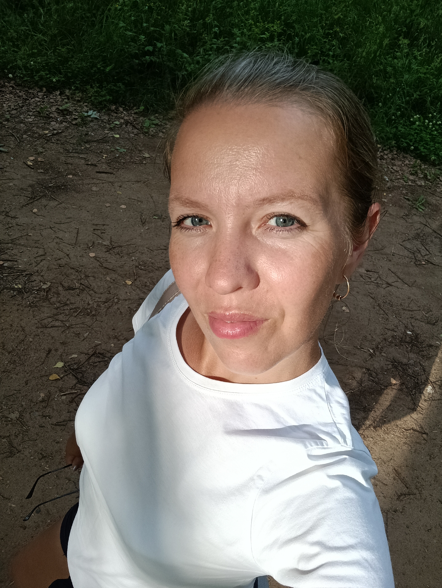 A woman is taking a selfie outdoors. She has fair skin, blonde hair with some gray streaks pulled back from her face, and blue eyes. She is wearing a white t-shirt and a gold hoop earring in her visible ear. She is holding a pair of black-rimmed glasses.
The background shows a dirt path with some fallen leaves and twigs, bordered by lush green foliage. The lighting suggests it is daytime, possibly late afternoon, due to the shadows. The overall scene appears to be a park or wooded area. The woman's expression is neutral, with her lips slightly parted. The image is taken from a high-angle perspective. There is no visible text in the image.