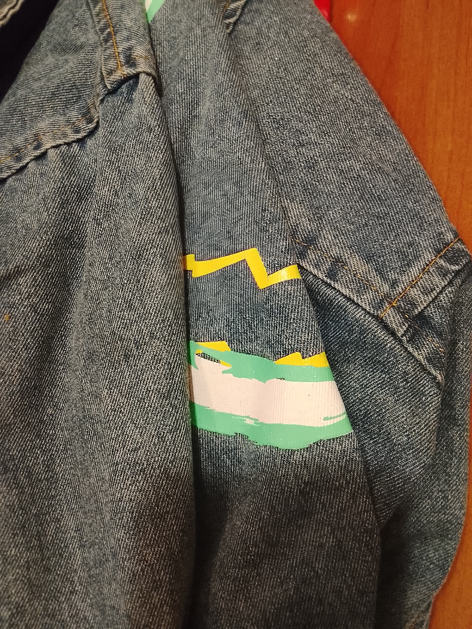 The image shows a close-up of a denim jacket sleeve with a painted design. The design consists of jagged yellow lines above a band of white with streaks of green paint. The jacket is hanging against a wooden surface. There is no indication of the city of Almaty, Kazakhstan.