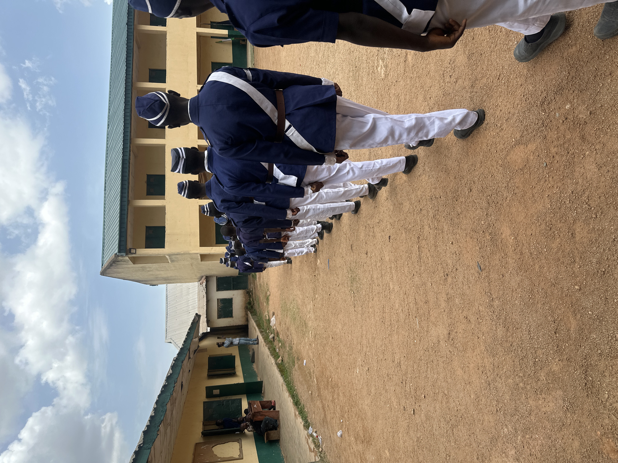 A line of young men wearing dark blue jackets, white trousers, dark shoes, and caps with white stripes stand at attention outdoors on a dirt ground in front of buildings. The buildings have yellow walls, green trim, and multiple windows. Some people sit or stand along the side of one of the buildings. The scene takes place under a partly cloudy sky. Given the building structure and the uniforms, this image is likely taken at a school or training facility in the city of Jos, Nigeria.