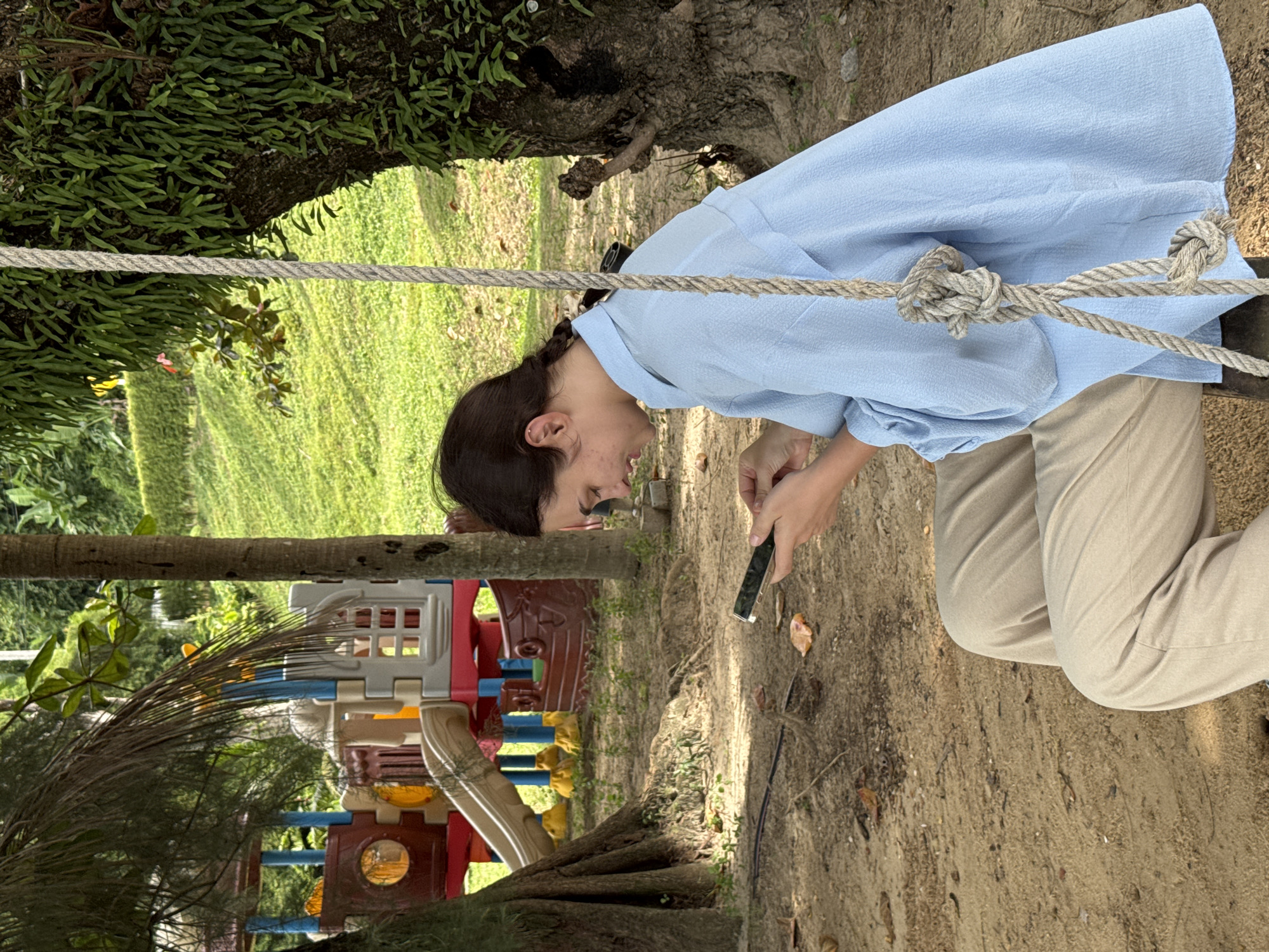 In Pa Khlok, Thailand, a young woman with dark hair pulled back in a braid sits on a rope swing. She is wearing a light blue, long-sleeved collared shirt and tan pants. The woman is looking down and appears to be holding and using a smartphone. She is positioned in the foreground of the image, with a sandy area and fallen leaves on the ground. In the background, a colorful playground structure is visible, with lush green grass and trees surrounding it. The rope of the swing is prominent in the frame, secured to a large tree with abundant green foliage. The overall scene suggests a casual, outdoor setting.