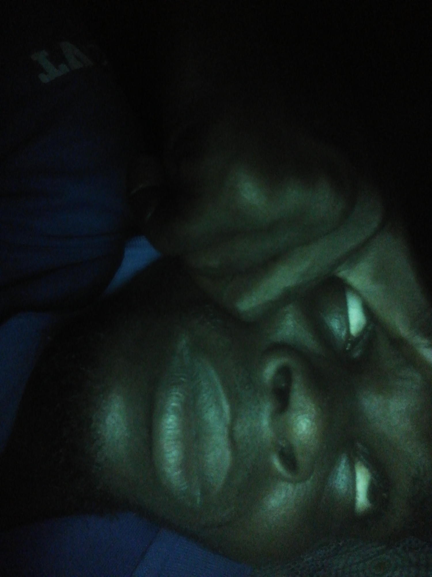 A close-up, low-angle shot of a person's face and part of their upper body lying on a surface or bed. The person has dark skin and a shirt with partial text visible in the upper left, appearing to read "LANT." The lighting is dim, casting shadows on the subject's face and surroundings. The environment is indistinct due to the lack of light. The city of Unknown, Unknown is not mentioned and cannot be inferred from the provided image.
