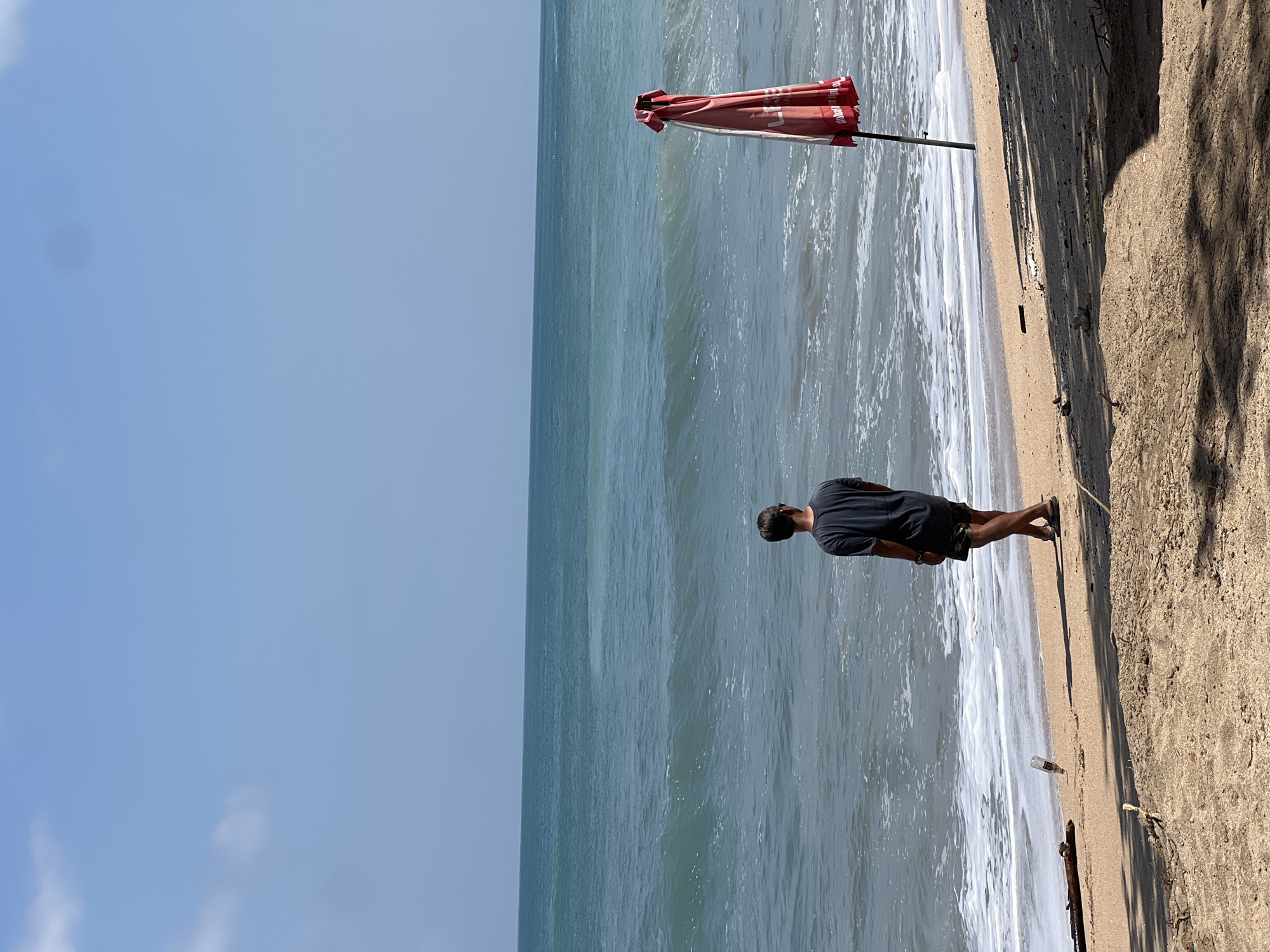 A young person stands on a sandy beach in Kuta, Indonesia, facing the ocean. The person is wearing a dark t-shirt and patterned shorts, with their back to the camera. In the background, waves crash onto the shore under a clear blue sky. To the right, a red beach umbrella is propped up on the sand. The scene appears to be a serene beach day with no other people visible in the immediate vicinity.
