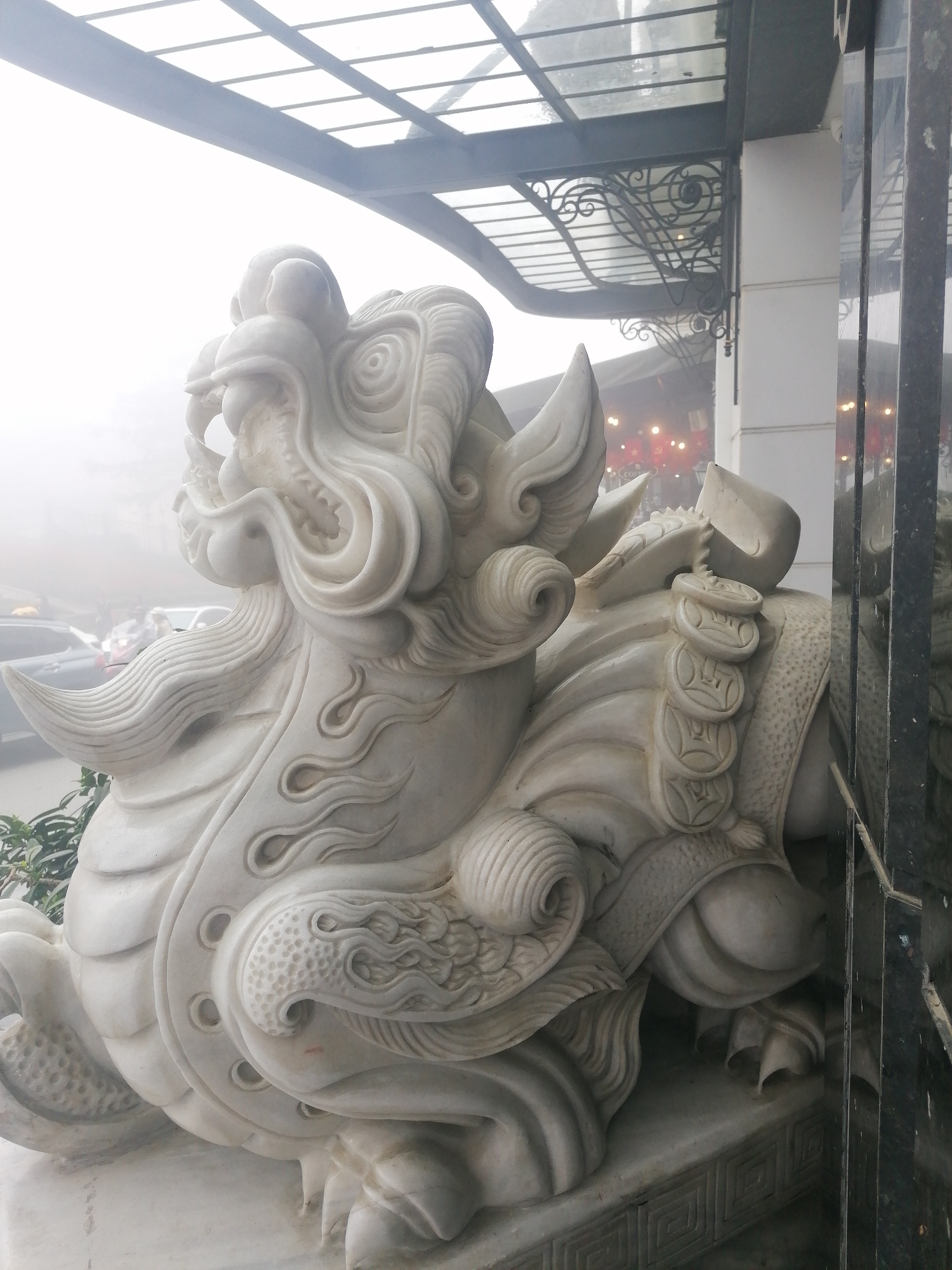 A large, white marble statue of a mythical creature, likely a Pixiu or Qilin, dominates the foreground, viewed from a slightly low angle. The statue is intricately carved, depicting a powerful beast with a fierce, ornate head featuring swirling mane-like elements, prominent fangs, and bulging eyes. Its body is adorned with scales, flame-like patterns, and a band of what appear to be ancient Chinese coins or medallions across its back. The creature rests on a decorative base with a geometric pattern.
In the mid-ground, a modern building with a dark metal and glass canopy or awning is visible. The canopy features elaborate, wrought-iron supports with decorative scrollwork.
The background is heavily obscured by a dense fog or mist, creating a soft, diffused light and significantly reducing visibility. Through the haze, indistinct shapes of parked cars and possibly other structures or lights can be vaguely perceived. Some green foliage is visible at the very bottom left corner, suggesting an outdoor setting.
The image captures a static scene. The weather is misty or foggy, giving the scene a serene and somewhat mysterious atmosphere, despite being daytime. The detailed craftsmanship of the statue and the architectural elements suggest an East Asian cultural influence, consistent with the provided location context of Sa Pa Ward, Vietnam. There is no clearly visible, readable text in the image.