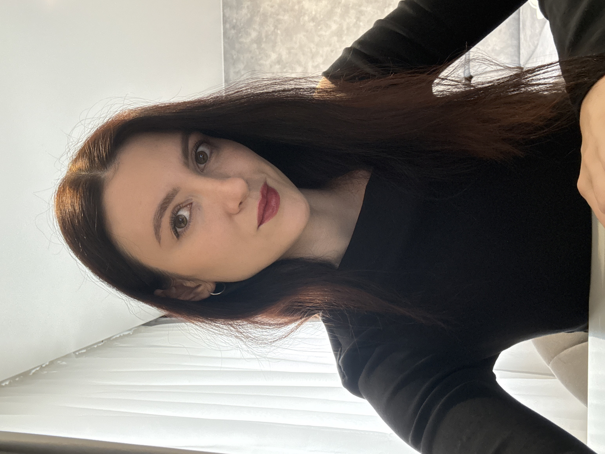 A rotated portrait features a young adult woman indoors in Kazan, Russia. She has long, dark brown hair, dark brown eyes, and is wearing dark red lipstick. Her expression is neutral as she looks directly towards the camera. She is dressed in a black long-sleeved top or dress, and a small silver hoop earring is visible on her left ear. The background consists of light-colored walls and vertical window blinds, indicating an indoor setting with natural light, likely from a window. A light-colored furniture piece, possibly a chair or couch, is partially visible in the bottom right corner of the frame. The image is captured at a steep angle, making the woman's head appear on the left side of the frame as if the photo was taken with the camera significantly tilted. No text is visible.