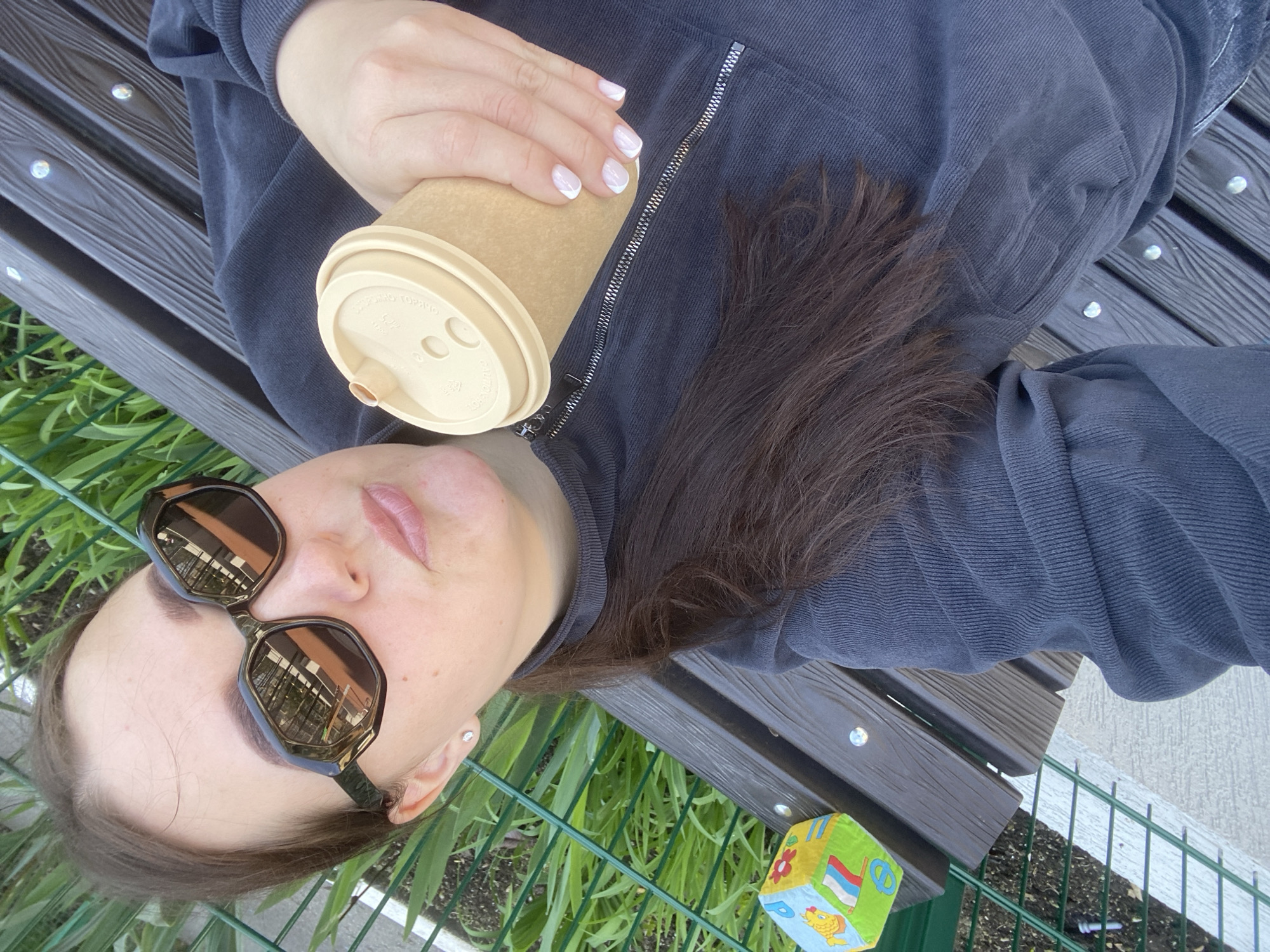 A woman lies on a park bench in Sochi, Russia. She wears sunglasses and a dark-colored, possibly corduroy, zip-up jacket. She holds a takeaway coffee cup in her hand. A child's block rests on the bench near green foliage and a fence.