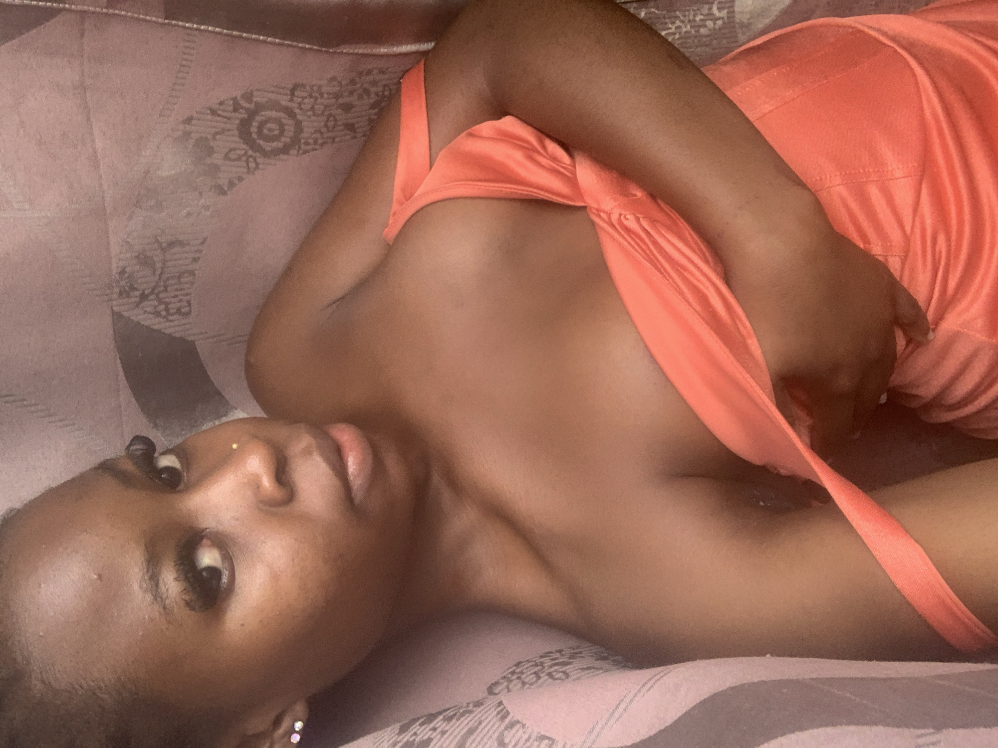 The image shows a close-up, overhead view of a young woman lying down, wearing an orange, off-the-shoulder dress. She has dark skin, dark hair pulled back, and is wearing subtle makeup, including eyeliner and mascara. She is also wearing a small nose stud and diamond stud earrings. The woman is positioned on a textured, pinkish-brown surface, possibly a sofa or bed, with a patterned fabric visible in the background. Her body is turned to her left, and she is looking towards the camera with a neutral expression. The lighting is soft, and the focus is primarily on the woman's face and upper body. There is no visible indication of the city of Akobo, Nigeria, within the image itself.