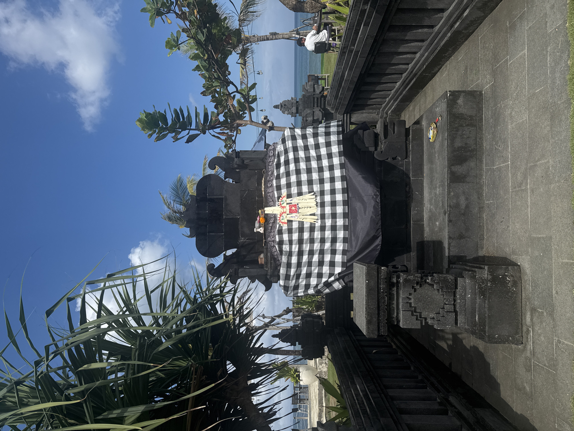 An outdoor, eye-level, medium shot captures a Balinese temple structure adorned with a black and white checkered cloth and offerings. The temple is crafted from dark stone and features intricate carvings. To the left of the temple, a large palm-like tree with long, green fronds partially obscures the blue sky, which is dotted with white clouds. To the right of the temple, a person in a white shirt and black hat sits at a table outdoors, facing away from the camera and towards the ocean. The ocean is visible in the background, with a table and chairs set up near the water. The ground in front of the temple is paved with large, gray stone tiles.