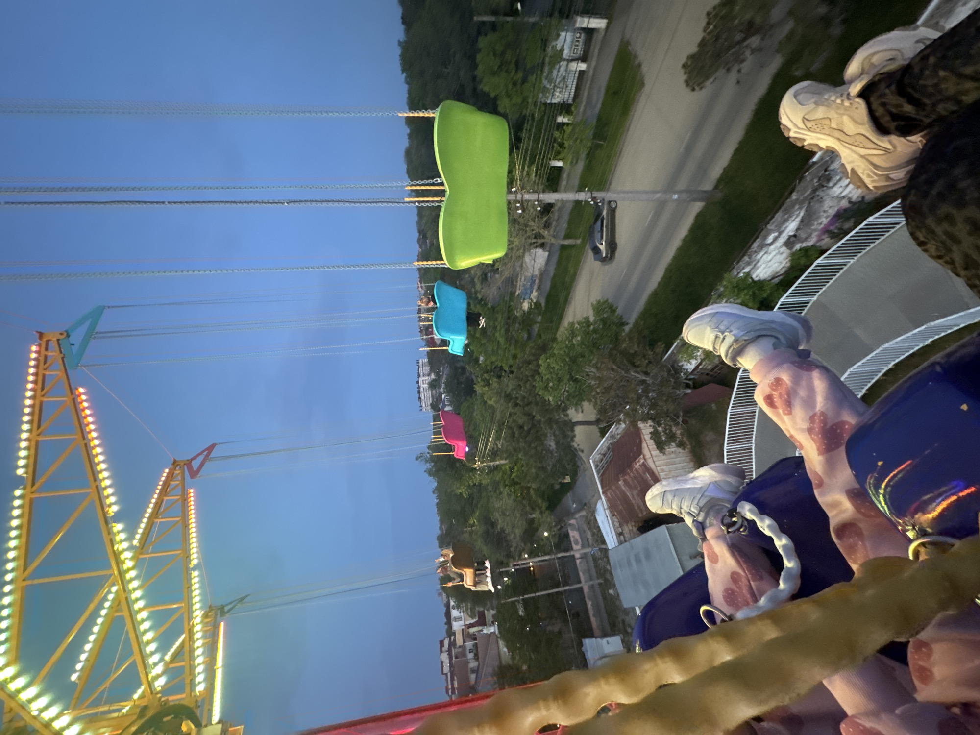 The image shows a nighttime view from a swing ride, likely a carousel or similar amusement park attraction. The perspective is looking down and out from a seat in the ride. Several other swings with different colored seats (green, blue, pink, and beige) are visible, suspended by chains. In the background is a road, trees, and buildings. The ride structure is on the left side, illuminated with yellow lights. The sky is a dark blue, suggesting it is dusk or early evening. People's feet and legs are visible in the foreground, suggesting occupancy of the seats within this swing ride. The location cannot be determined.