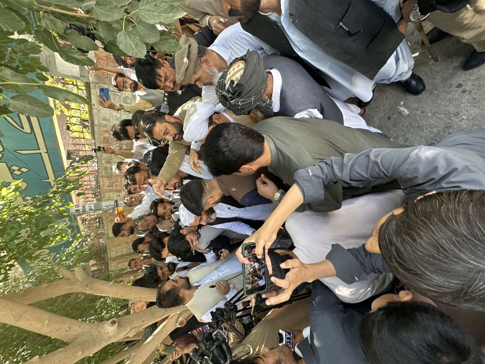 In Quetta City Tehsil, Pakistan, a dense crowd surrounds a person speaking into a microphone. Many individuals in the crowd are holding up smartphones, seemingly recording or photographing the speaker. The crowd consists primarily of men, many wearing traditional clothing, including turbans. Professional broadcast cameras are visible near the speaker. The setting appears to be outdoors, next to a tree.