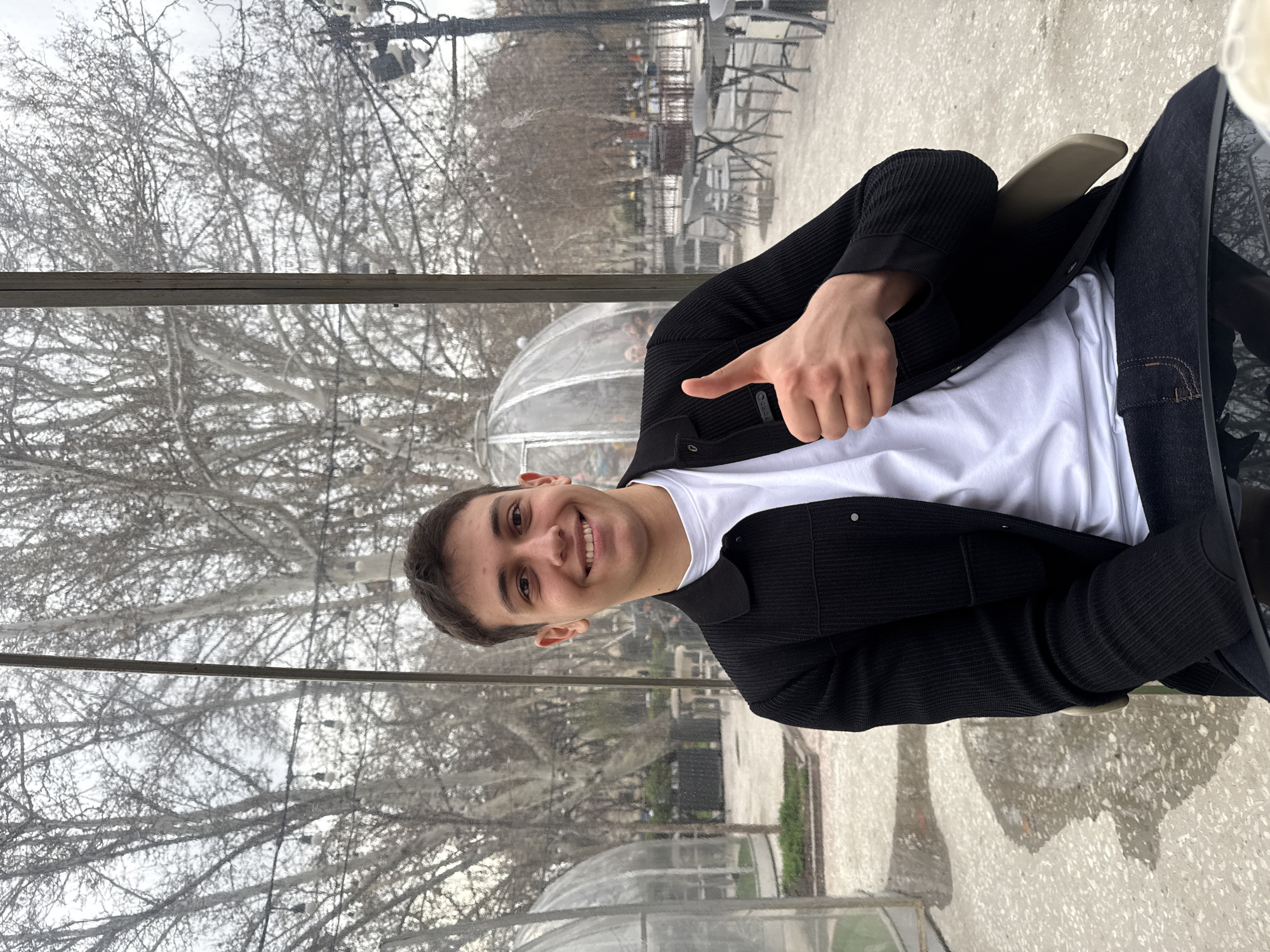 A young man is shown in the image, seated outdoors and giving a thumbs-up. He is wearing a white t-shirt under a black sweater or jacket, and dark jeans or pants. The background indicates he is in an urban park-like setting in Yerevan, Armenia. Behind him, there are bare trees and semi-transparent, dome-shaped structures, likely outdoor dining areas, characteristic of establishments in the city. A metal frame is visible in the foreground, possibly part of the enclosure or structure where he is seated. He is sitting on a chair, and the ground appears to be paved.