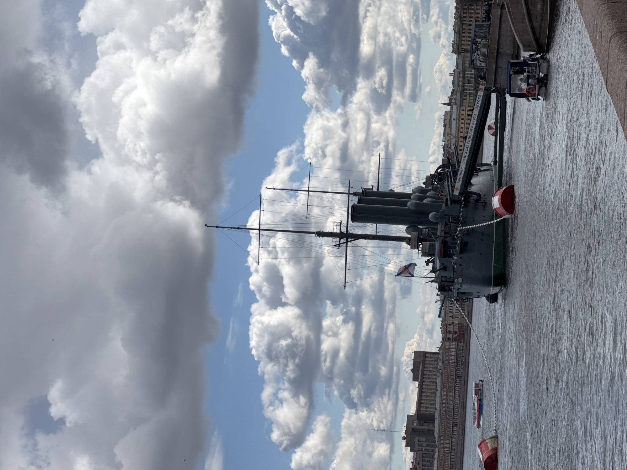 The image depicts the Cruiser Aurora, a Russian warship museum, located in Saint Petersburg, Russia. The warship is anchored in the Neva River, with its stern towards the viewer. The vessel's masts and four smokestacks are visible against a backdrop of a partly cloudy sky. The river is choppy, reflecting the overcast sky. Buildings line the riverbank on the right side of the image. A small boat with passengers is docked near the warship, and mooring buoys are visible in the water. A gangway extends from the ship to the dock.