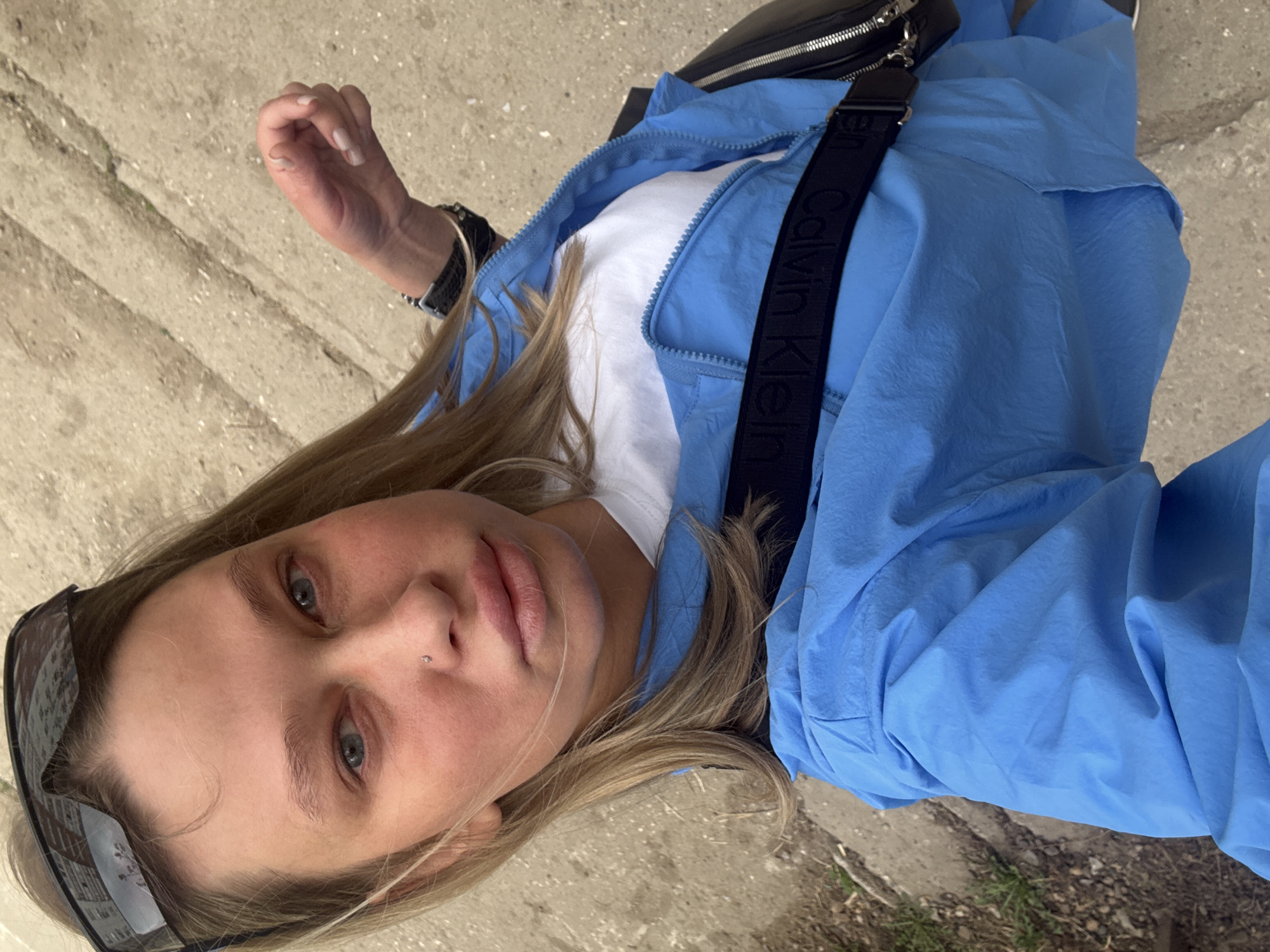 A young adult female is shown in a close-up, top-down view, dressed in a white t-shirt and a light blue zip-up jacket, with a black Calvin Klein crossbody bag. Sunglasses are partially visible on the left side of the frame. She has blonde hair and fair skin, and a nose piercing. The background shows concrete pavement and some greenery, suggesting an outdoor location. The scene appears to be taken in an urban setting. It is not possible to determine that this is Perm, Russia.