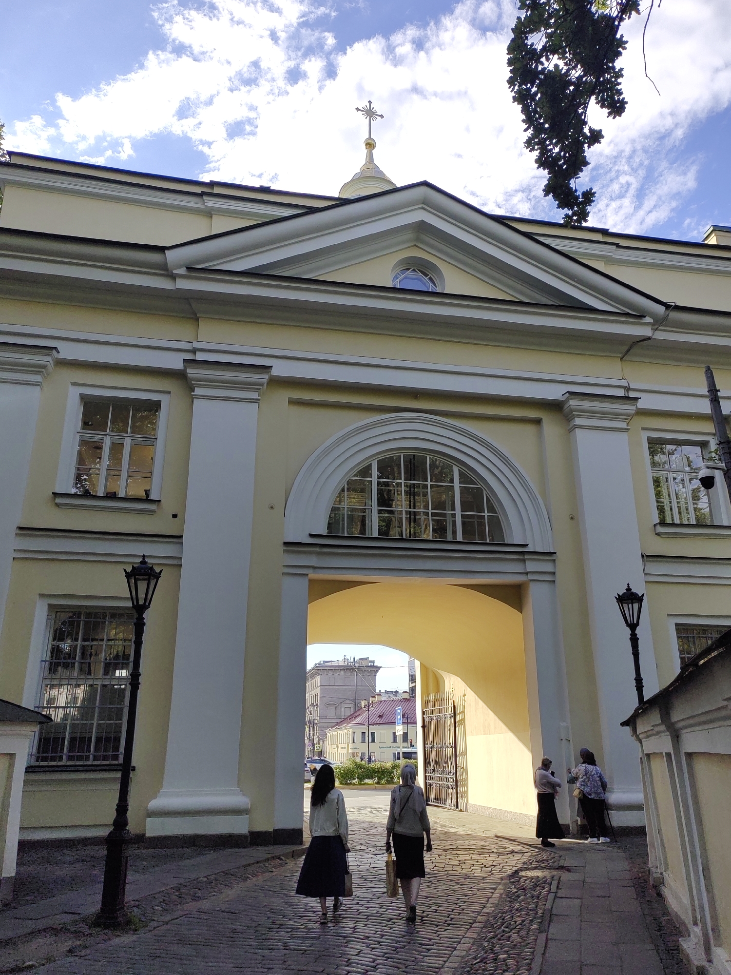 In this full outdoor, low-angle shot, two women walk away from the camera on a cobblestone path in front of a pale yellow, classical-style building. The building features a large arched entrance with a semi-circular window above it. On either side of the entrance, there are tall, white columns and windows with decorative frames. A black lamppost stands to the left of the entrance. The sky above is blue with scattered white clouds, and a tree branch with dark green leaves hangs down from the upper right corner. A cross on a golden dome is visible at the very top of the building. Through the arched entrance, a gate is visible, as well as a street with buildings and a car. The lighting suggests it is daytime, with shadows indicating the sun is to the left. The scene appears to be a peaceful, historical setting in Saint Petersburg, Russia.
