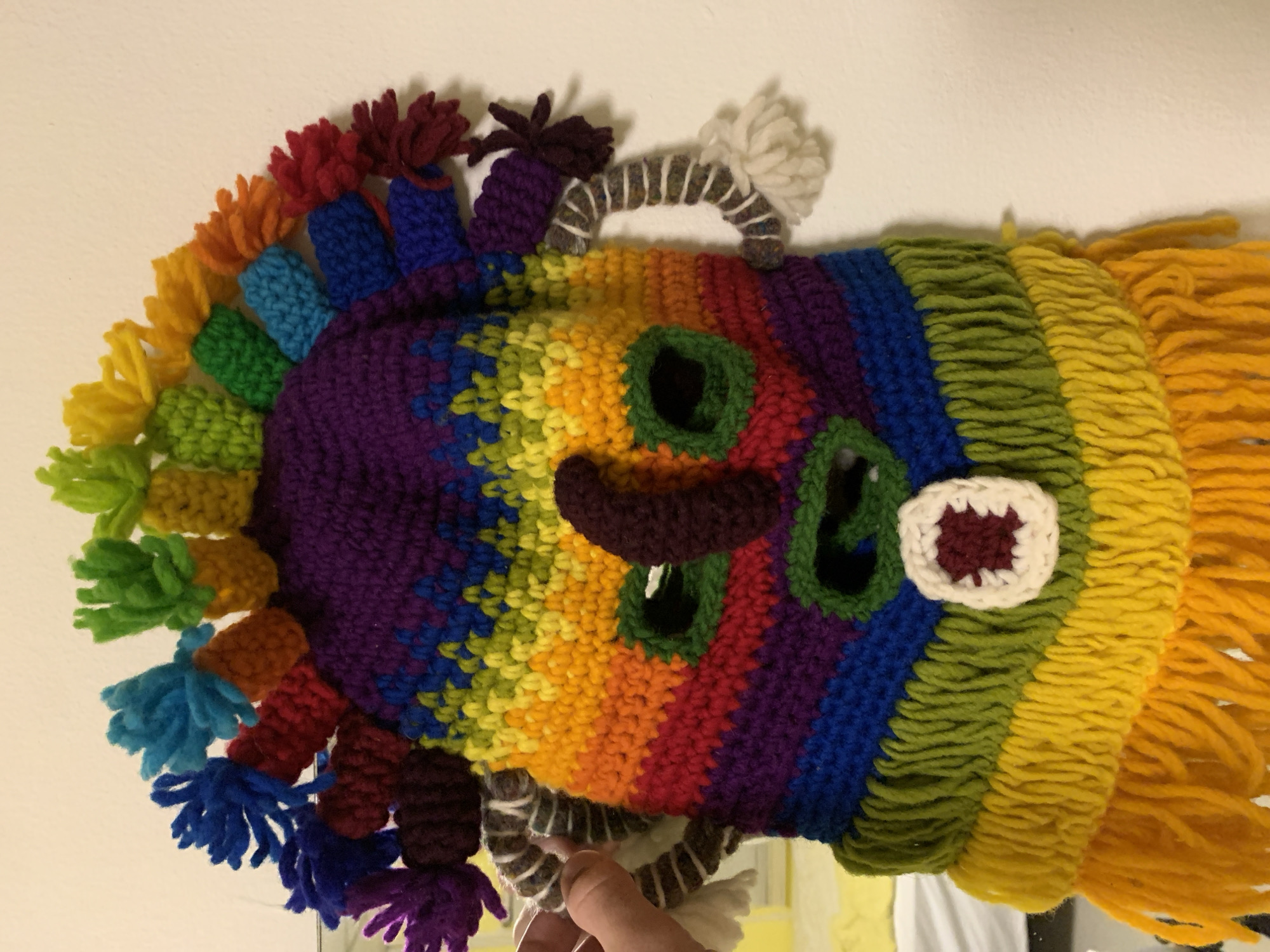 An unidentifiable person's hand is holding a colorful, crocheted mask that is hanging on a white wall. The mask is shaped like a face and is adorned with multiple yarn tassels in a rainbow of colors, resembling a headdress. The mask has a purple crocheted top, a rainbow striped face with green eye openings, and a small white and burgundy square detail below the mouth opening. The mask also has two ear-like appendages on either side, made of twisted gray and white yarn. The background is a plain white wall, with a sliver of a yellow object visible in the lower left corner of the frame, possibly a bed or a piece of furniture. The lighting in the image suggests an indoor setting, possibly a home.