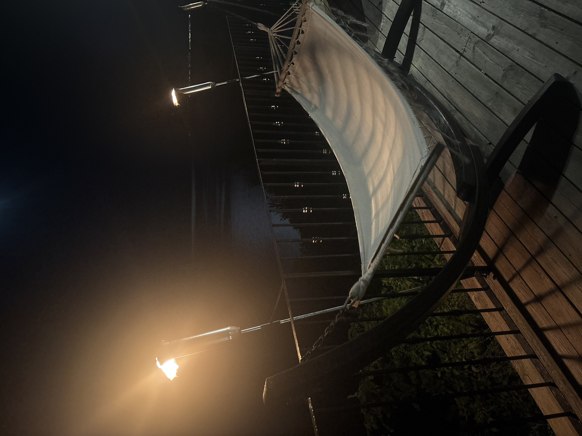 A nighttime shot captures a hammock on a wooden deck. Two tiki torches illuminate the scene with warm light. The hammock is made of white fabric and is suspended from a dark wooden stand. A metal railing separates the deck from a dark, wooded area below. In the background, a body of water is visible, with faint lights suggesting a distant city. The sky is dark, with a hint of moonlight.