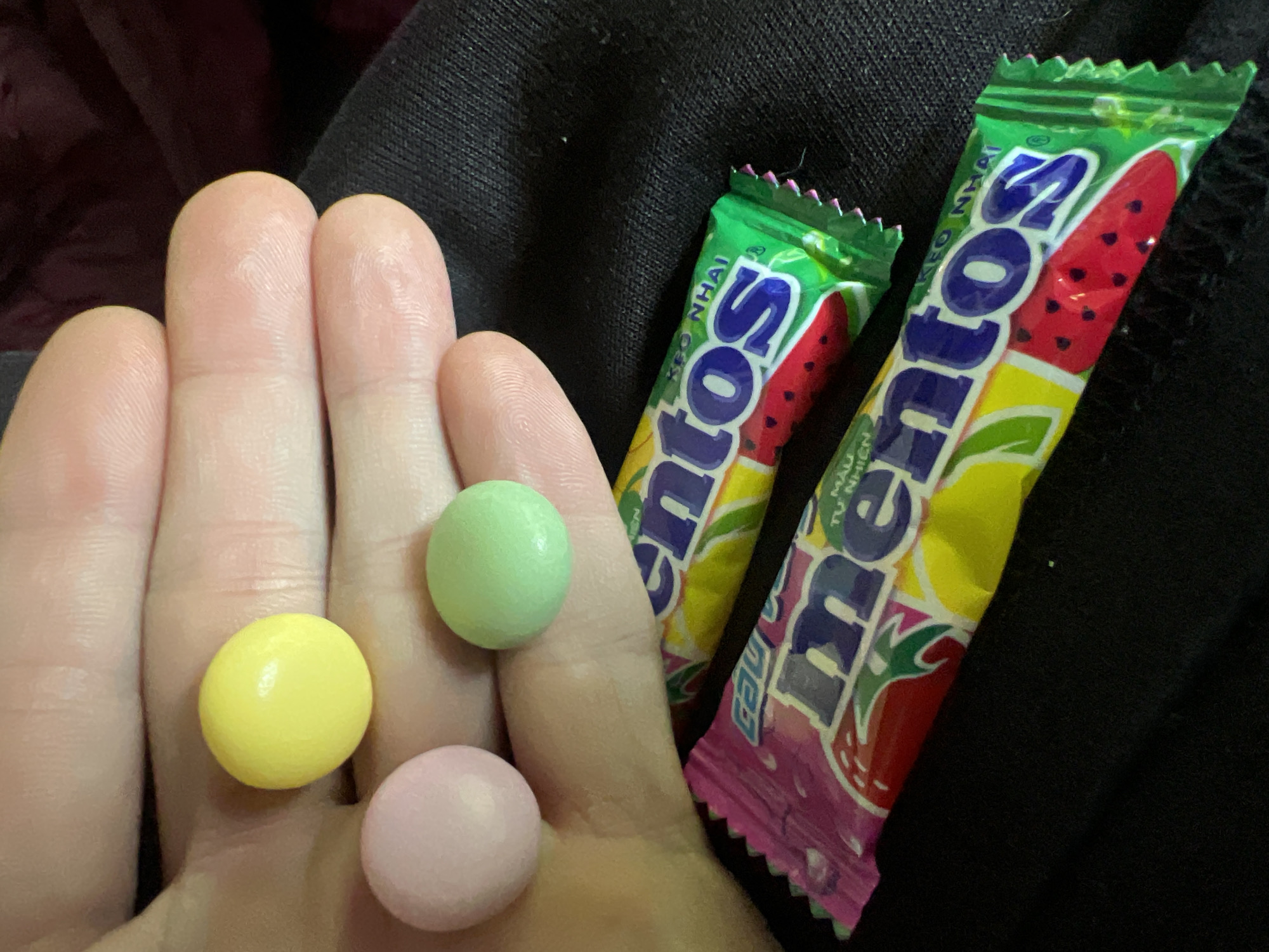 A close-up shot reveals a person's open left hand, holding three round candies - one yellow, one green, and one pink. Adjacent to the hand are two unopened packets of Mentos candies, showcasing watermelon flavors. The background consists of black clothing material. The location is inferred to be Đăk Hà, Vietnam based on the packet writing "KEO NHAI". The location is assumed based on the Mentos packaging.