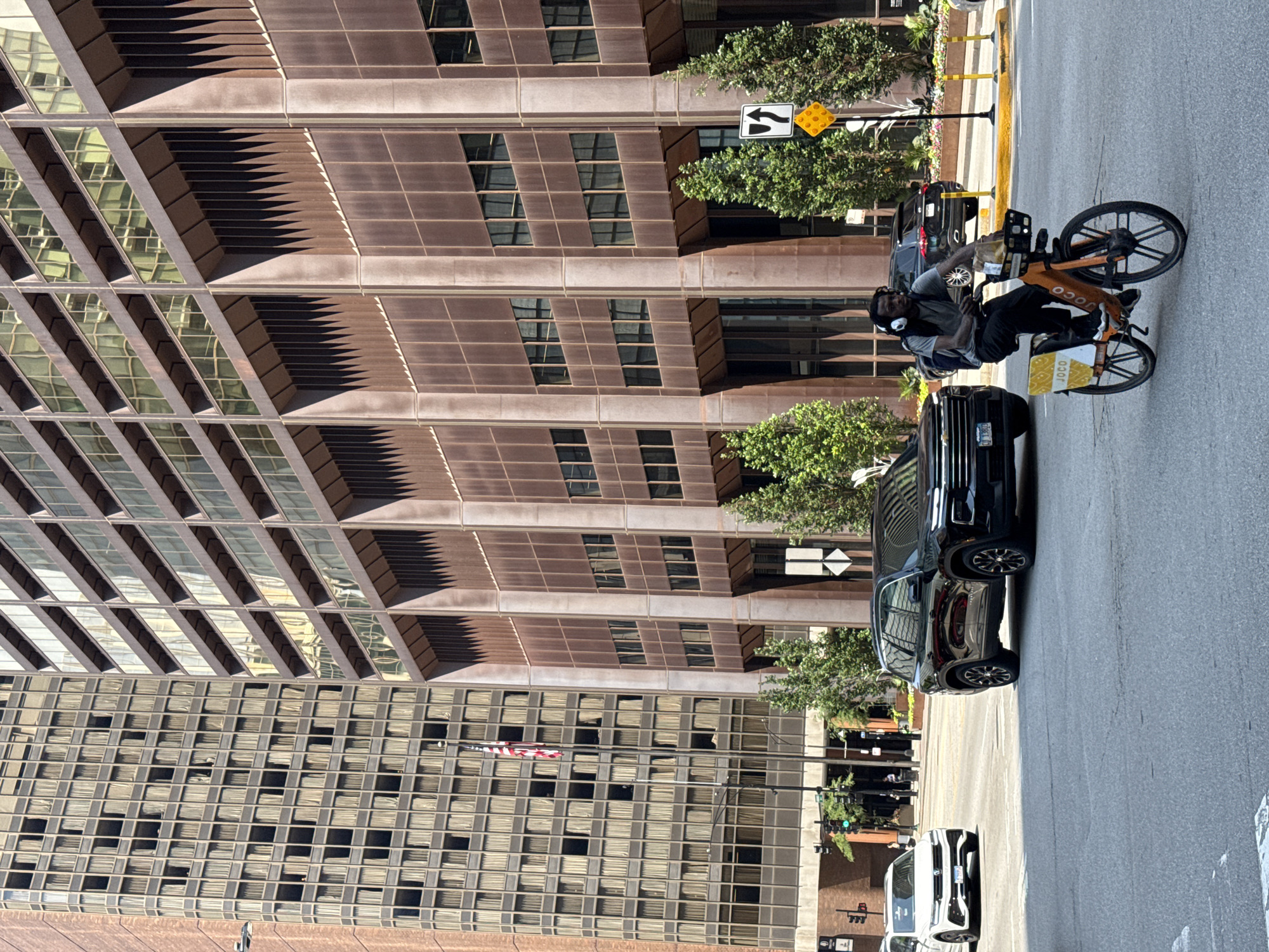 A man wearing headphones and a backpack is riding an orange bicycle with "JOCO" branding in white lettering on the side, across a city street. He is moving from left to right in the frame. In the background, a black SUV is parked on the right side of the street, and a white SUV is driving away on the left side of the street. Behind the vehicles are tall, modern buildings with many windows. An American flag is visible on a flagpole in front of one of the buildings on the left. The scene appears to be a daytime street in Chicago, Illinois. Traffic signals and street signs are also present.
