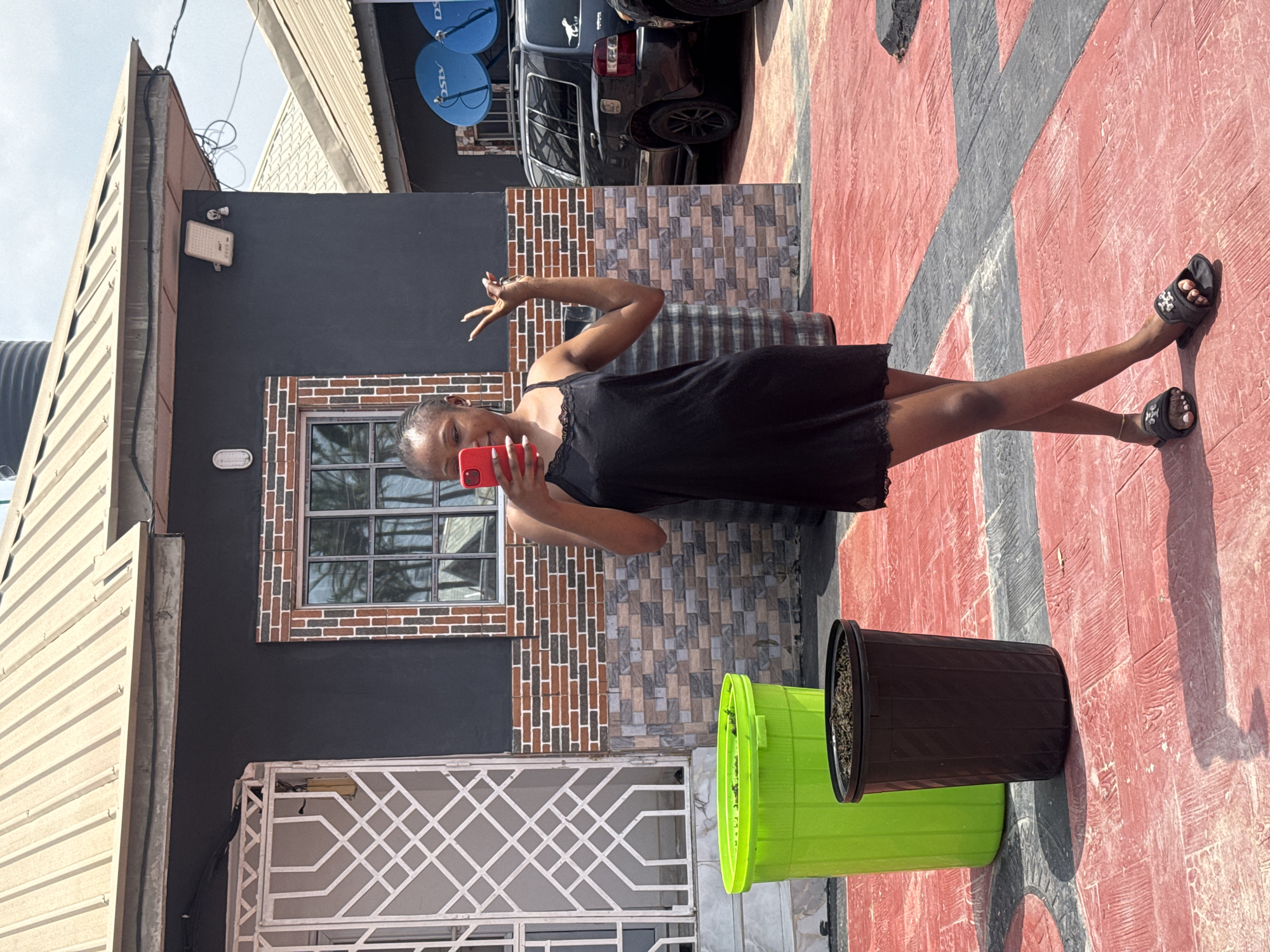 A young woman, dressed in a black slip dress, stands outdoors and takes a selfie with a red smartphone. She is positioned in front of a building with a black facade and a brick-patterned tile accent around a window. Her right arm is raised, with her hand making a peace sign towards the camera. She is wearing black sandals with white embellishments. The ground in front of her is a paved surface with a red and grey geometric pattern. To her right are two large plastic planters, one lime green and the other black, both containing soil. In the background, a black SUV is visible, along with two blue satellite dishes mounted on the building. The lighting suggests it is daytime, and the scene appears to be in a residential or commercial area, consistent with a location context of Abuja, Nigeria. The woman's pose and gesture suggest a casual and perhaps cheerful disposition.