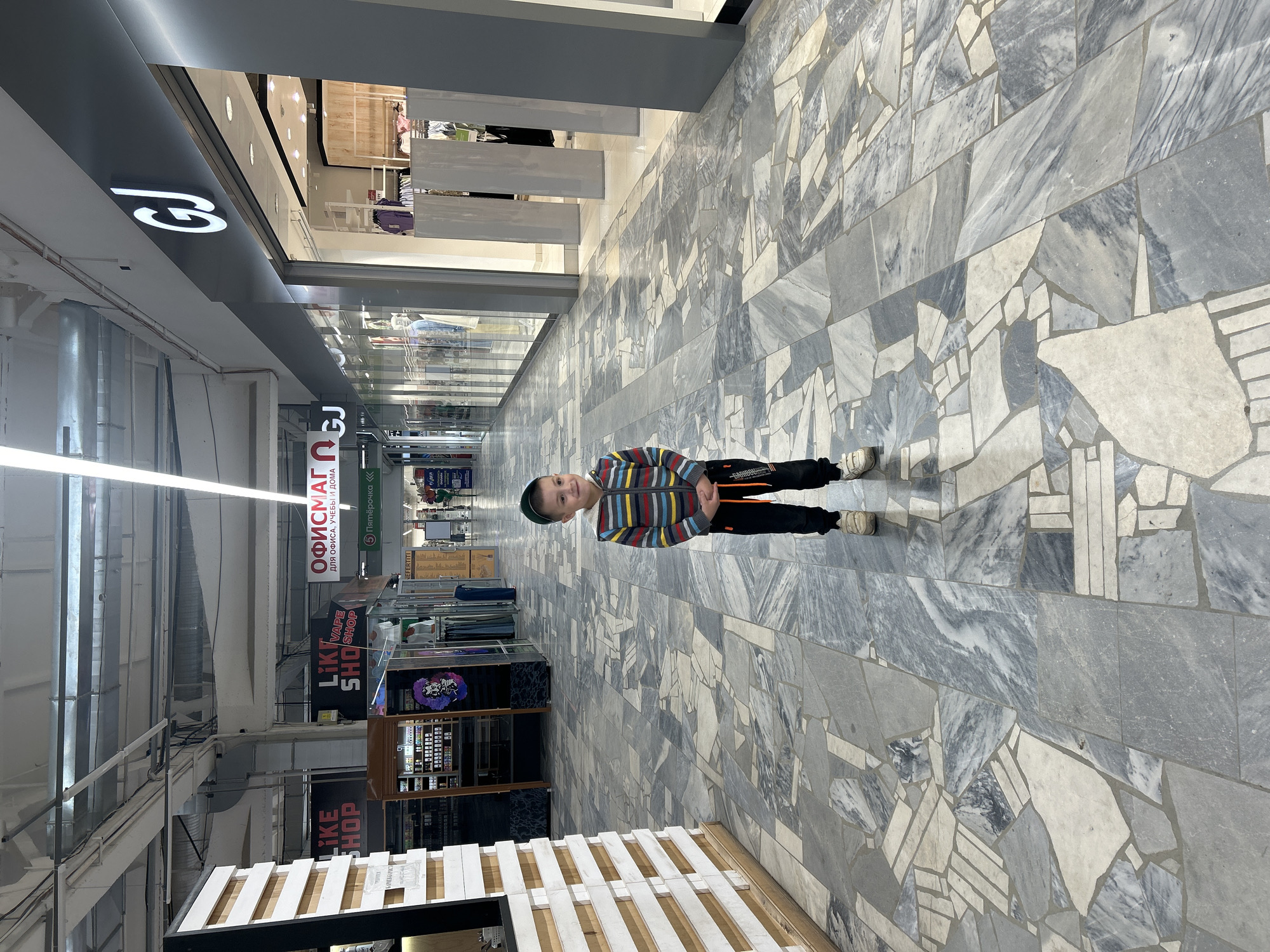 The image depicts an indoor shopping mall corridor with a young boy standing in the center. He is wearing a striped hooded jacket, black pants, and white sneakers. The boy is looking towards the left side of the frame with a slight smile.
The mall corridor has a unique floor design composed of irregularly shaped marble tiles in shades of grey and white. On the left, there are storefronts with signs that read "LIKE SHOP" and "VAPE SHOP". Further down the corridor, there is a sign for "ОФИСМАГ" which translates to "OFFICEMAG" and is followed by text that says "for office, study and home". On the right, there is a prominent store with a large "GJ" logo. The ceiling has exposed ductwork and bright fluorescent lighting.
The image appears to be taken in the city of Chaikovsky, Russia, based on the signage in Russian. The overall environment suggests a typical indoor shopping center.