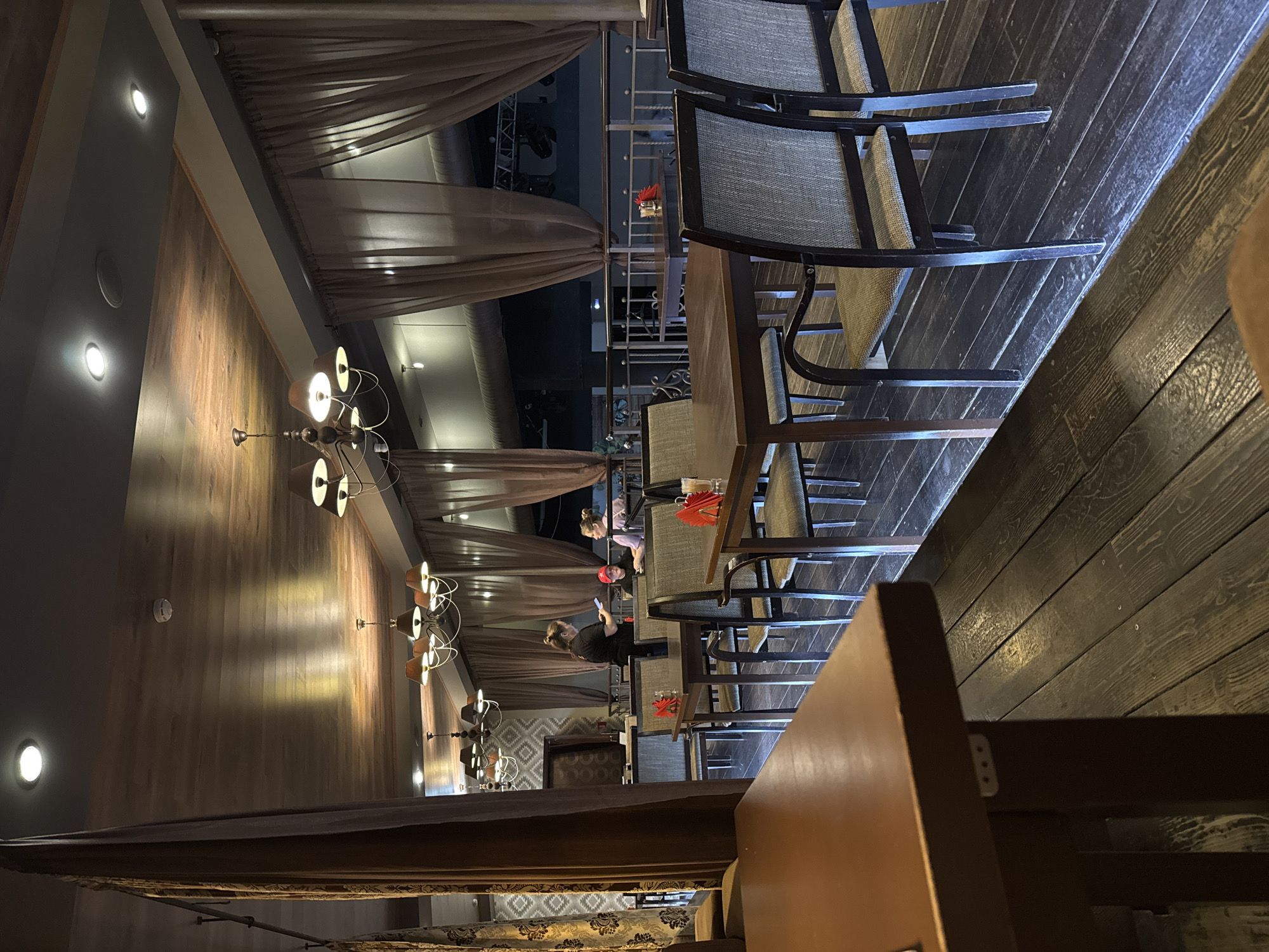 The image depicts the interior of a restaurant in Balezino, Russia. The dining area is lit by chandeliers and recessed ceiling lights, creating a warm ambiance. The walls are paneled with wood. The room is furnished with wooden tables and chairs with woven seats. Some tables are occupied by patrons. There are brown drapes along the side of the room. A metal railing is visible outside, beyond the drapes.