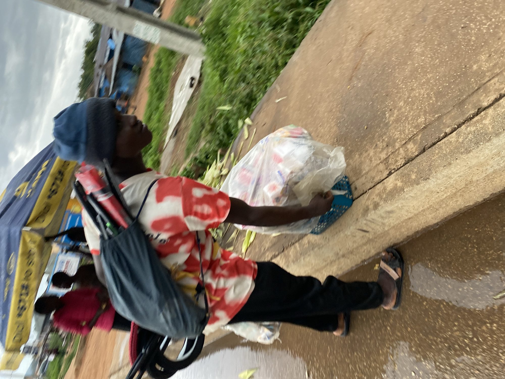 The image, taken in Bukuru, Nigeria, shows a person standing or walking alongside a wet roadside. The individual, likely male, is dressed in a blue knitted beanie, a red and white tie-dye patterned t-shirt, black pants, and dark sandals. They are carrying a black shoulder bag filled with several stick-like objects, possibly umbrellas, and are holding or standing next to a transparent plastic bag containing various items, suggesting merchandise, and a blue basket.
The ground is visibly wet with puddles, indicating recent rainfall, and the sky is overcast, suggesting either a cloudy day, early morning, or late afternoon. In the blurred background to the left, another person in a pink top is partially visible near what appears to be a blue and yellow canopy or umbrella, possibly part of a market stall, which has some indistinct text printed on it. Beyond the concrete edge of the road, green vegetation and simple structures can be seen. The scene depicts a street vendor engaged in commerce in what appears to be a semi-urban or rural setting.