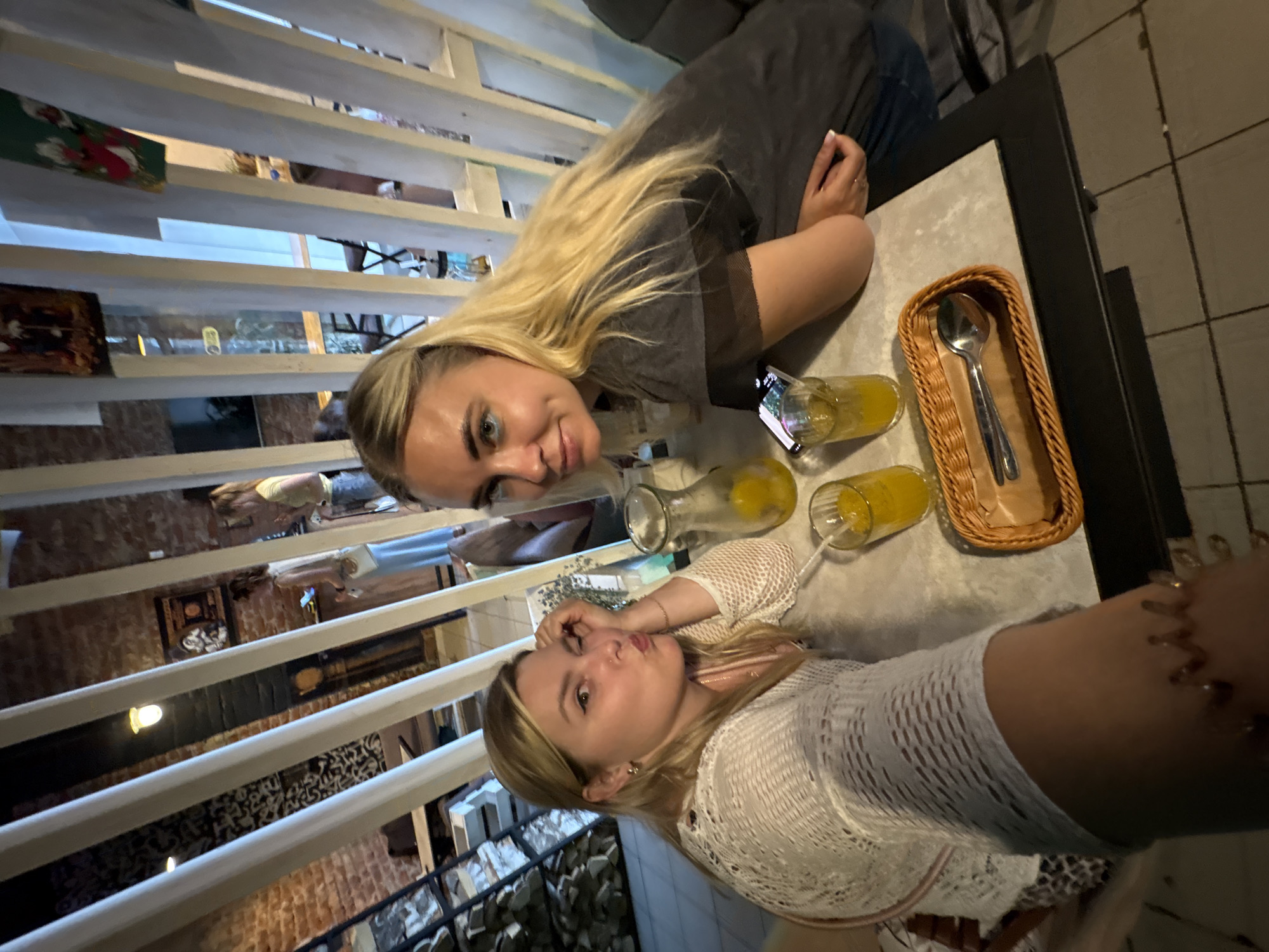 Two young women are seated at a table in what appears to be a cafe or restaurant. The woman on the left is pouting and has her hand on her chin, wearing a white open-knit sweater. The woman on the right has blonde hair and is wearing a dark t-shirt. They are both looking towards the camera. On the table in front of them are two glasses of orange liquid, a pitcher of the same liquid, and a small wicker basket containing a spoon. The environment is dimly lit with exposed brick walls and white wooden partitions. In the background, other patrons can be seen, along with shelves and decor. The scene is consistent with a casual dining establishment. The presence of items like a pitcher of drinks and glasses suggests they are enjoying refreshments.