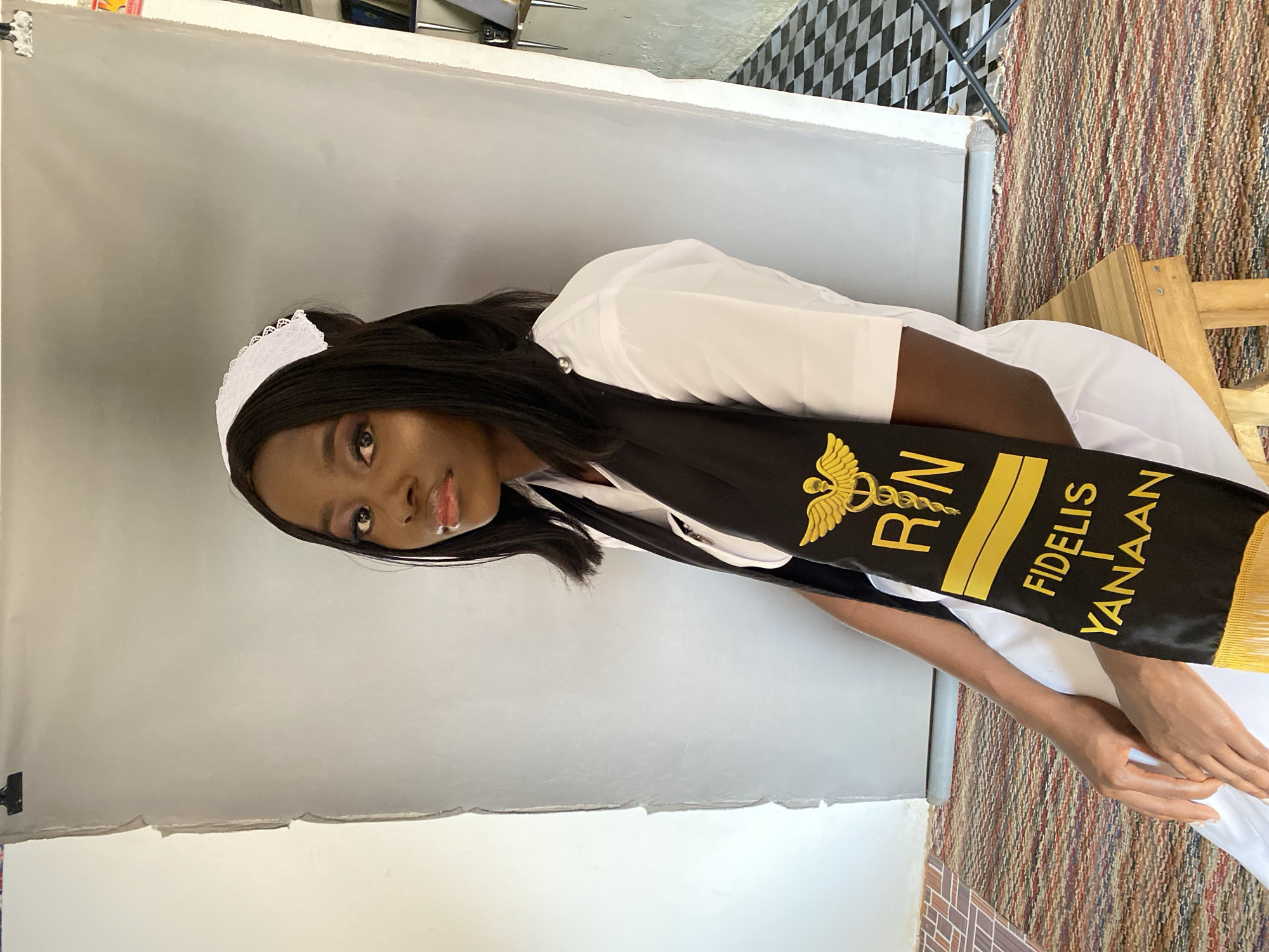 This is a portrait of a young woman, likely a recent nursing graduate, photographed indoors. She has a dark complexion and long, dark hair, and is looking directly at the camera with a gentle, proud expression.
She is wearing a white nursing uniform, consisting of a white top or dress and a small, white cap with a delicate lace edge. Draped over her shoulders is a black stole adorned with prominent gold embroidery. The embroidery features a caduceus symbol, the letters "R N" (Registered Nurse), and the text "FIDELIS I YANAAAN". Her hands are positioned in front of her, either clasped or resting.
The background consists primarily of a light gray or beige seamless backdrop, characteristic of a professional photography setting. To the right, the floor transitions from a multi-colored striped woven rug to a black and white checkered pattern. A simple wooden stool or table is also visible on the right side of the frame. The lighting is bright and even, suggesting either studio lighting or abundant natural light during daytime. The image conveys a sense of accomplishment and celebration, likely marking her graduation or professional certification in Vom, Nigeria.
