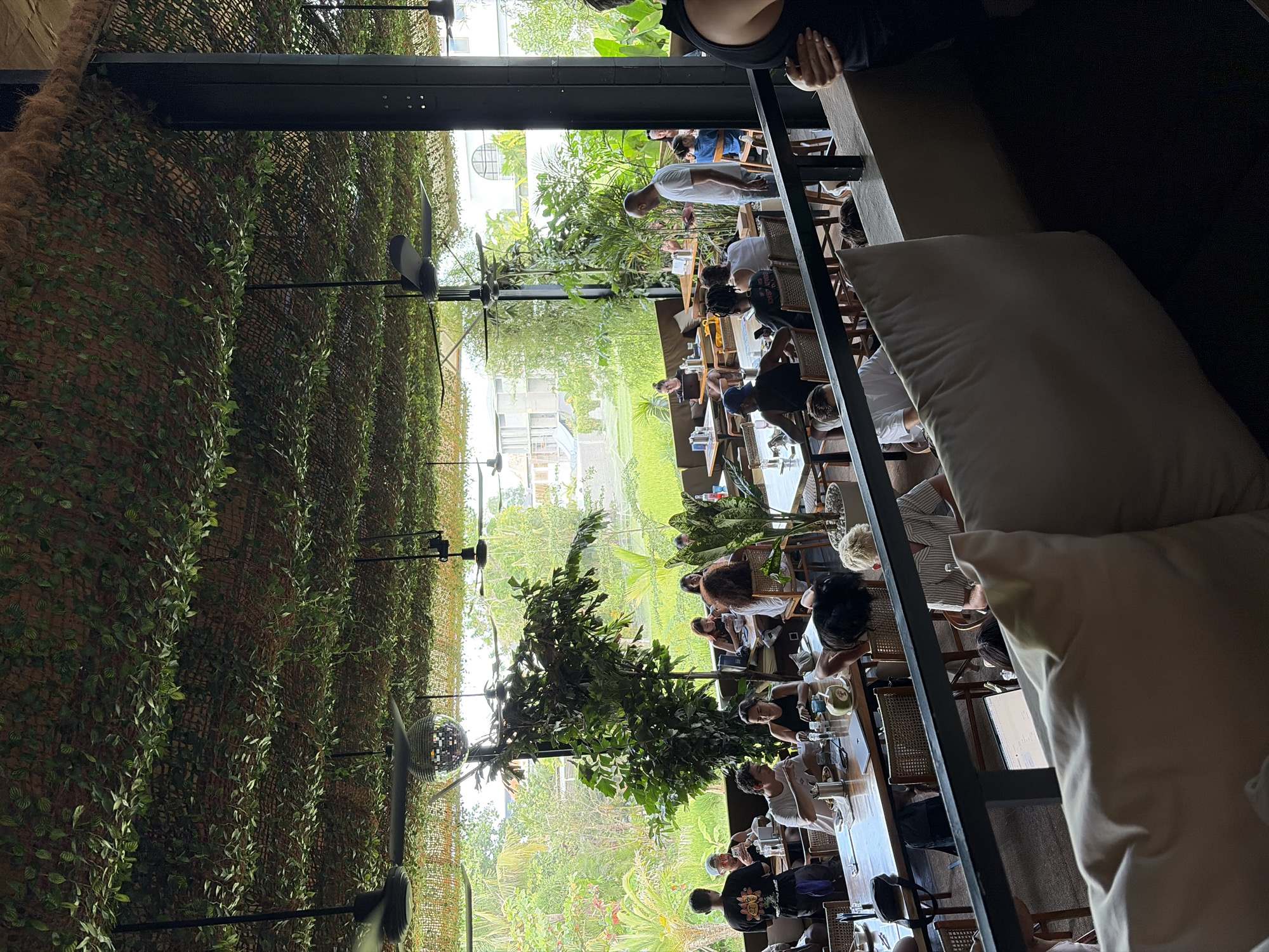 The image depicts a bustling restaurant interior with an outdoor seating area, likely in Mengwi, Indonesia, given the lush green tropical vegetation and what appears to be rice paddies visible through the open-air structure. The ceiling is covered with a mesh netting adorned with faux greenery, creating a naturalistic ambiance. Multiple ceiling fans are visible, along with a disco ball hanging near the center of the space.
The restaurant is populated with numerous patrons seated at various tables, engaged in dining and conversation. Some individuals are interacting with their mobile devices, while others are actively eating. The furniture consists of wooden tables and rattan chairs. Plants are strategically placed throughout the dining area, enhancing the tropical theme. In the foreground, a large white pillow rests on a dark sofa, hinting at a more relaxed seating arrangement.
The lighting suggests it is daytime, with natural light filtering in from the outside. The overall atmosphere appears to be lively and casual, typical of a popular dining establishment in a tropical location. The view beyond the restaurant's confines reveals a verdant landscape, characteristic of the Indonesian countryside.