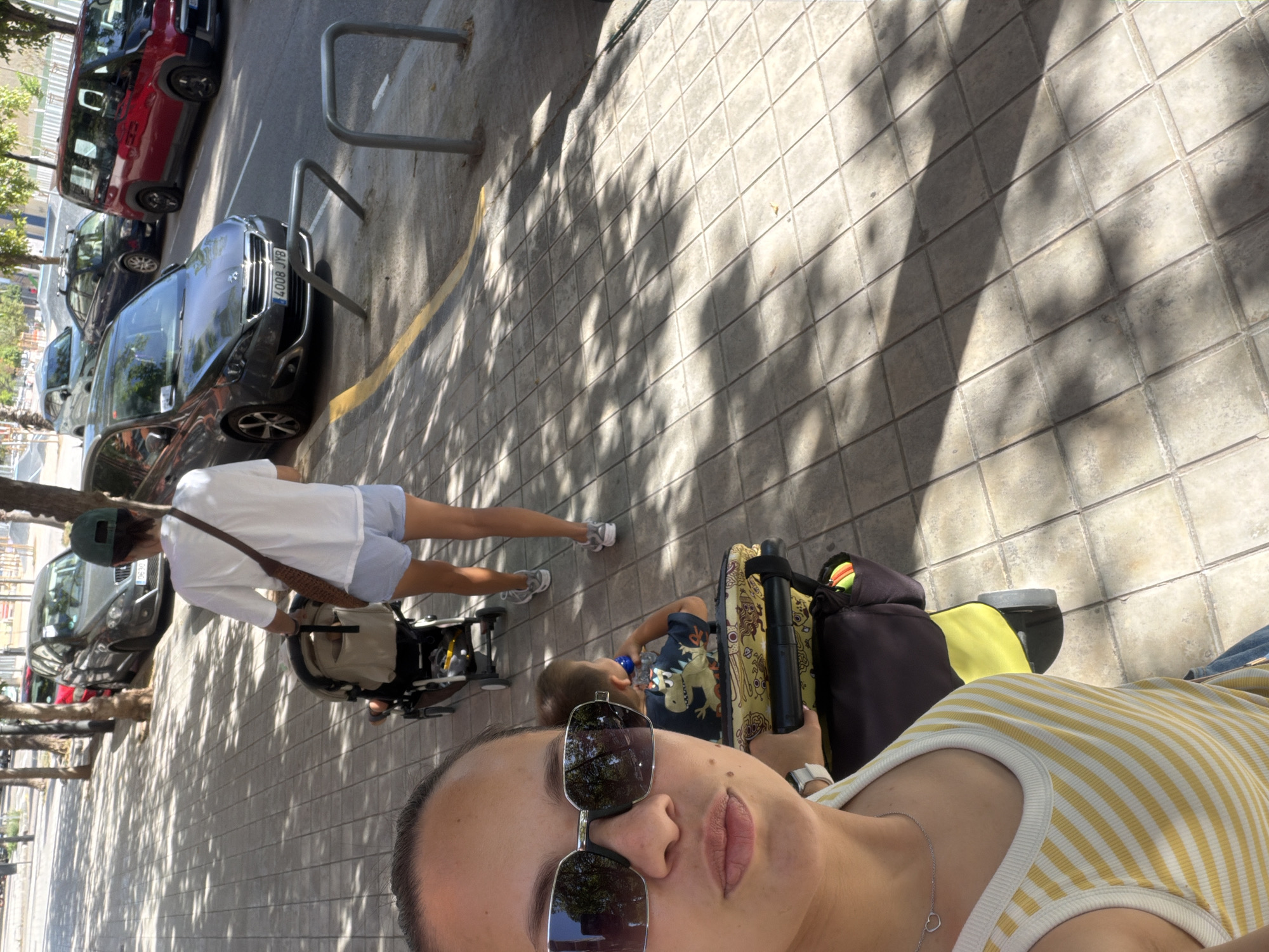 A selfie-style medium shot captures a woman in the foreground, wearing a yellow and white striped tank top and sunglasses, looking at the camera. To her right, a child sits in a stroller. Behind them, another person, possibly a man, pushes a stroller with a child in it. The scene takes place on a sun-dappled sidewalk lined with trees. Several cars are parked on the street to the right, and a red minivan is visible further down the road. The environment suggests an outdoor urban setting, likely a street in a city.