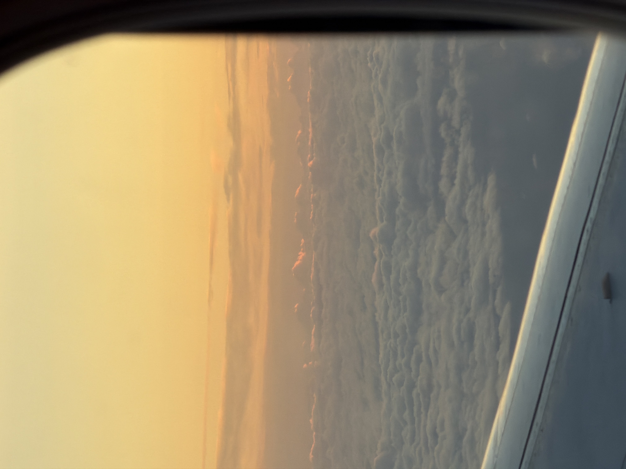 The image captures a view from an airplane window, looking out at a vast expanse of clouds during sunset. The sky is painted in warm hues of orange and yellow, with thin, wispy clouds scattered at various altitudes. Below, a thick layer of fluffy white clouds stretches as far as the eye can see. The lower portion of the image shows a part of the airplane's wing, its metallic surface reflecting the soft light of the setting sun. The view is framed by the dark border of the airplane window, suggesting the perspective of a passenger in flight. The scene is serene and atmospheric, conveying a sense of height and distance above the earth. The location is specified as Emelyanovo Airport, Russia, though this cannot be directly confirmed from the visual information alone.